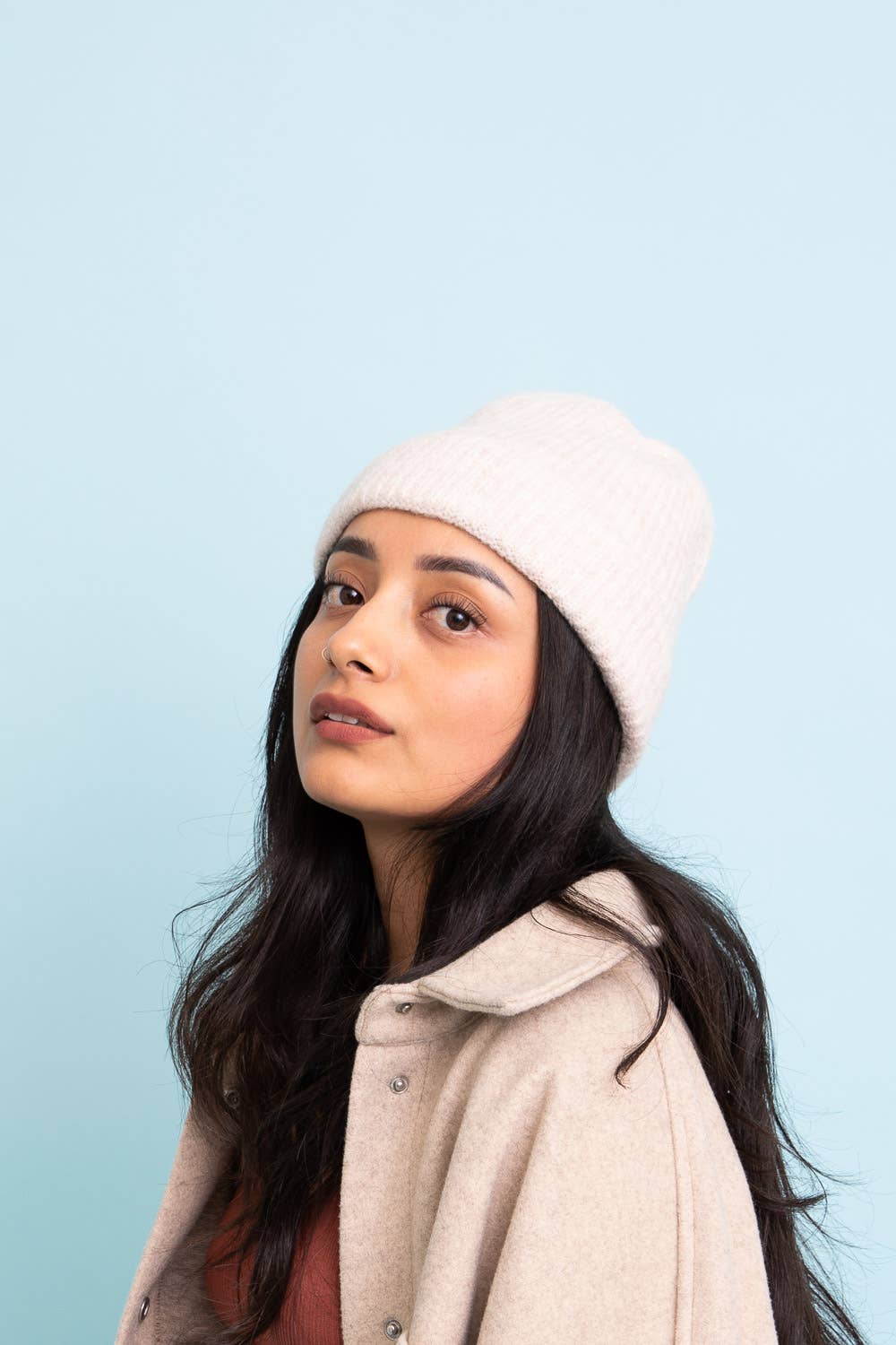 A woman with long dark hair wears the Versatile Cuffed Beanie by Leto Accessories—a cozy rib knit hat—in cream, paired with a light beige coat against a pale blue background. The perfect fall accessory.