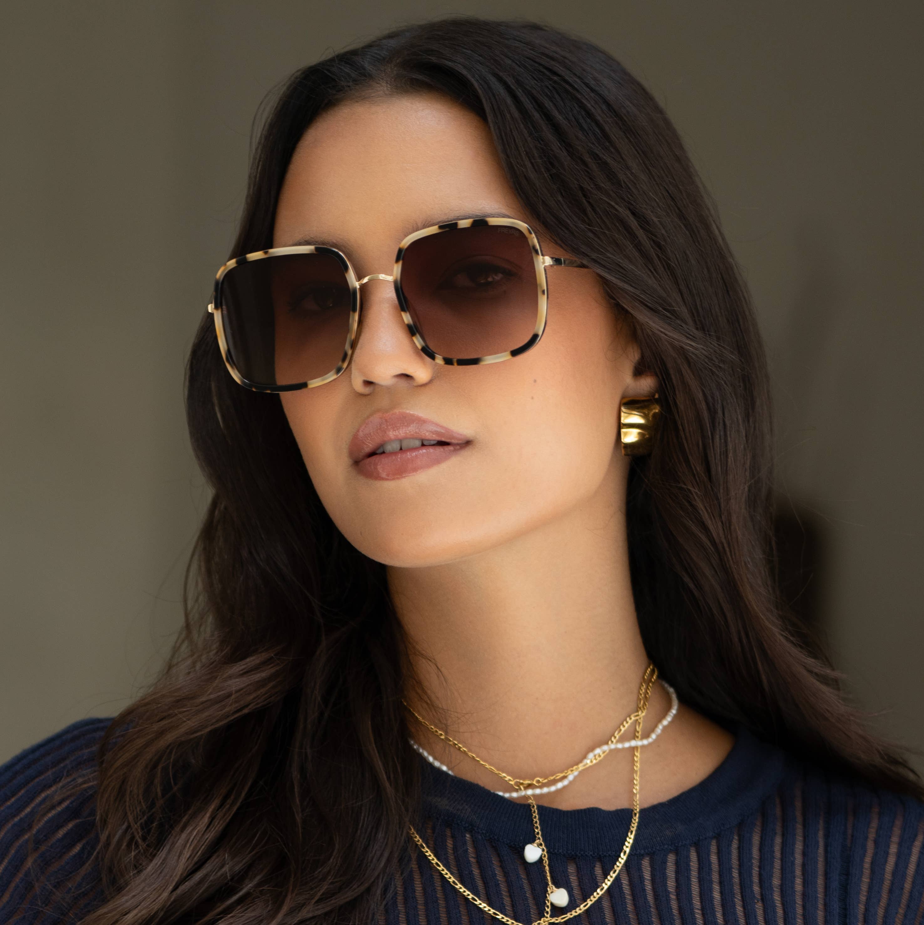 A woman with long dark hair wears FREYRS Eyewear Cosmo Acetate Womens Square Sunglasses, featuring handmade acetate frames in tortoiseshell. She pairs them with gold earrings, layered necklaces, and a navy blue sheer top, looking confidently at the camera.