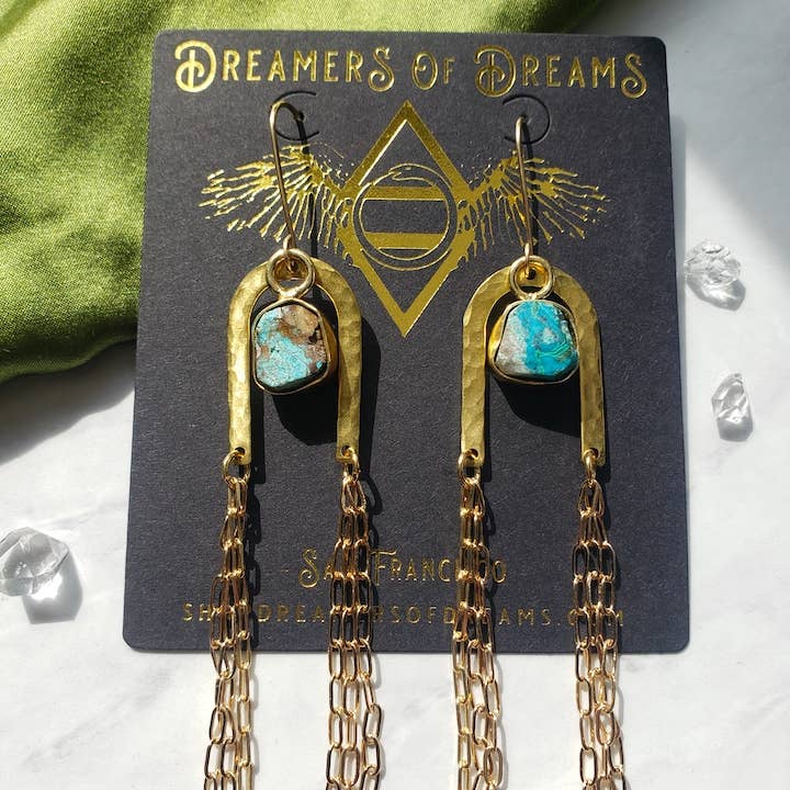 The Gold Rainbow Arch Chain Duster Earrings by Dreamers of Dreams feature turquoise stones and multiple gold chain dangles, displayed on a branded card with small clear crystals scattered nearby.