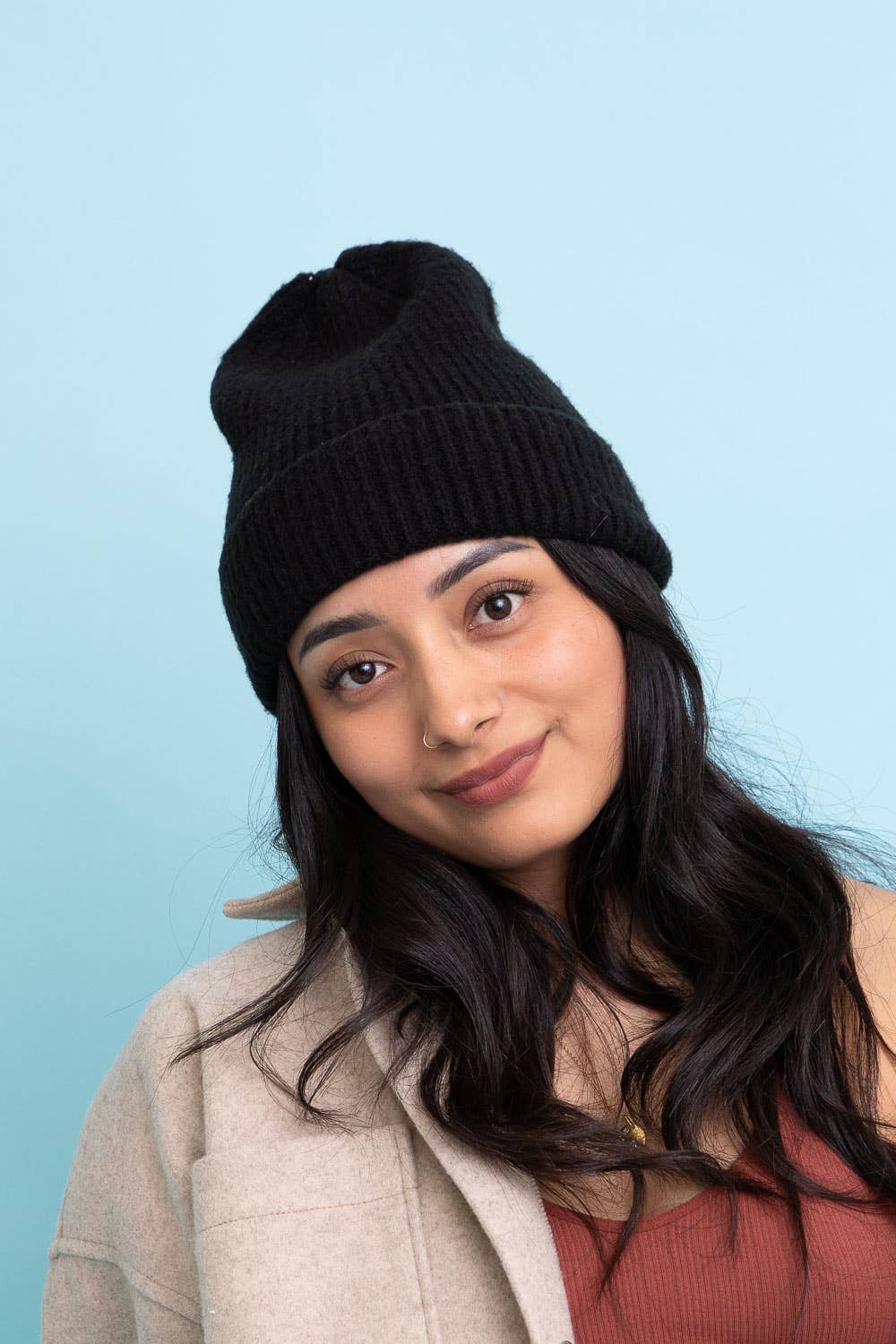 A young woman with long dark hair smiles softly against a light blue background, wearing the Versatile Cuffed Beanie – Cozy Rib Knit Hat by Leto Accessories, paired with a beige jacket and rust top—a chic fall look.