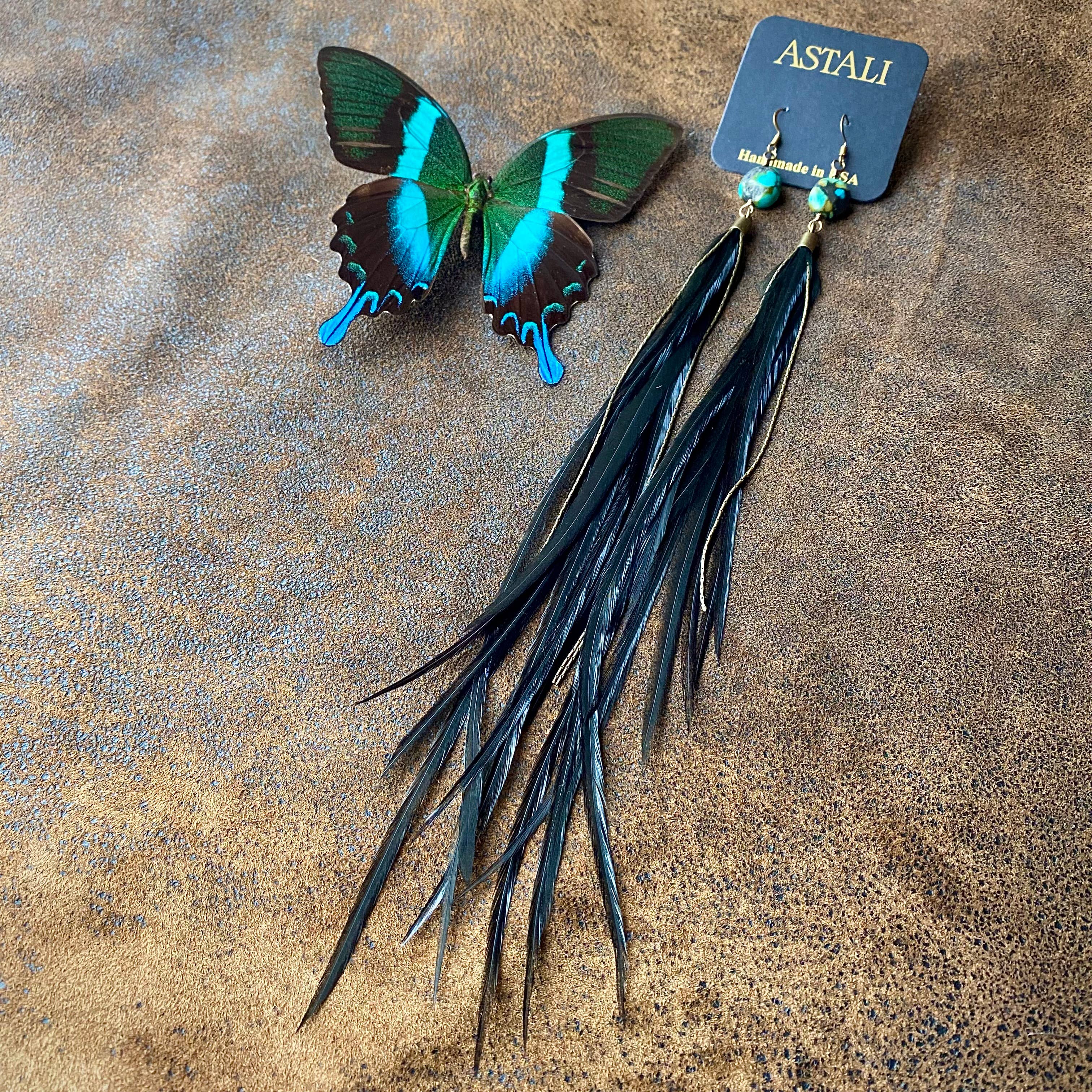 A pair of ASTALI Feather & Turquoise Statement Earrings, crafted with sustainable coque feathers and antique brass, rest beside a green and black butterfly on a brown textured surface.