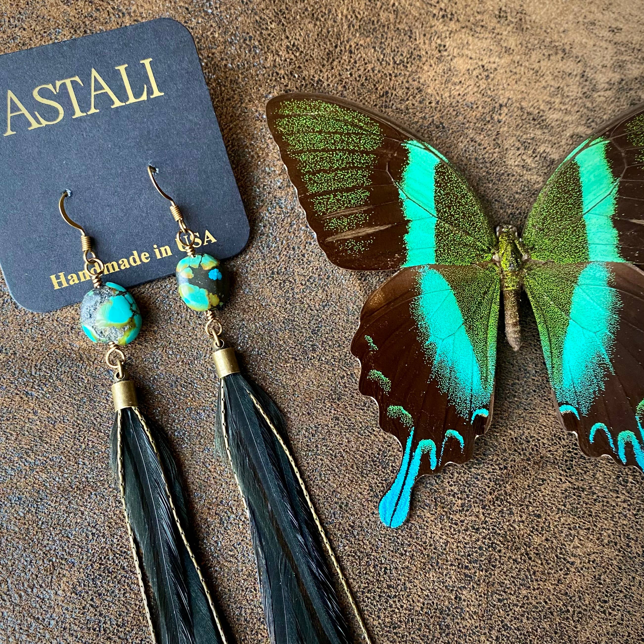 A pair of ASTALI Feather & Turquoise Statement Earrings—crafted with antique brass and sustainable coque feathers—shown on a branded card beside a vibrant green and brown butterfly on a textured brown surface.