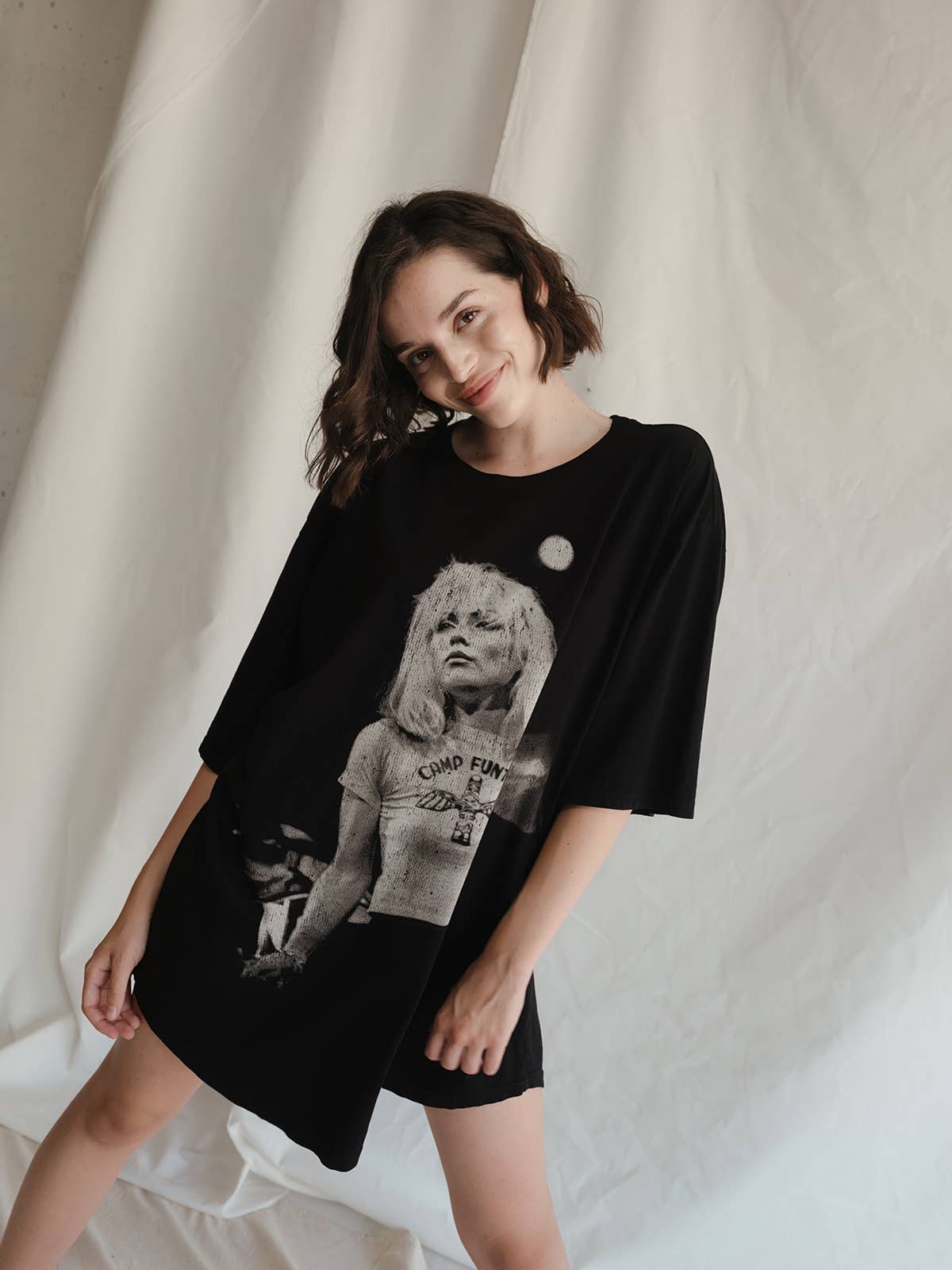 A woman with short brown hair smiles subtly while wearing the Blondie Camp Oversized Tee by People Of Leisure, an ethically made black T-shirt dress with a monochrome woman and moon graphic, standing before a white fabric backdrop.