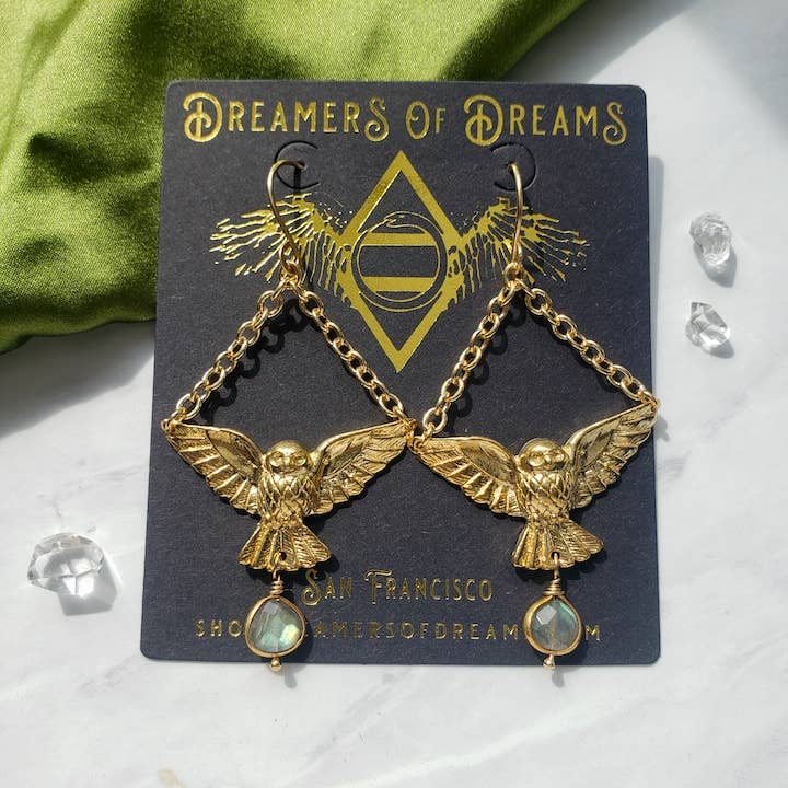 Gold Owl Labradorite Charm Dangle Earrings by Dreamers of Dreams—gold plated owls with outstretched wings and labradorite gems, shown on a branded black card beside green fabric and crystals.