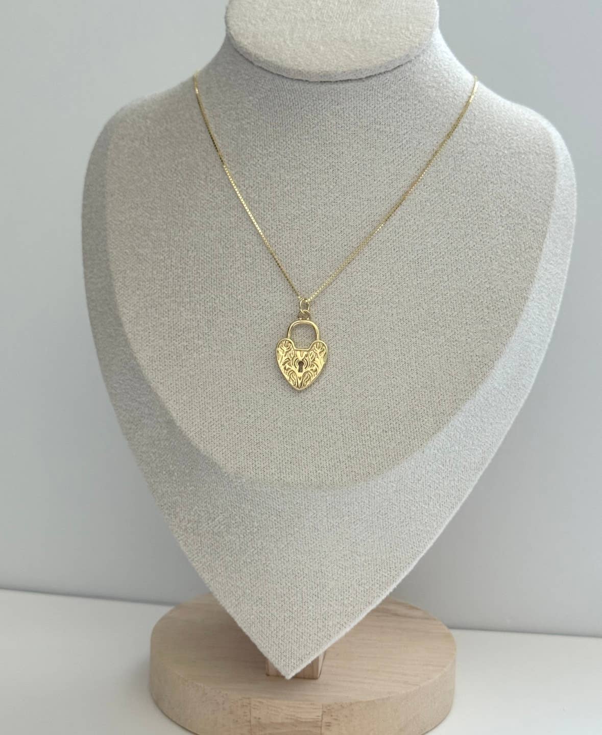 The Since Ive Been Loving You Necklace by Tramps+Thieves features a gold vermeil heart-shaped pendant with an engraved floral design, shown on a light gray jewelry stand with a wooden base.