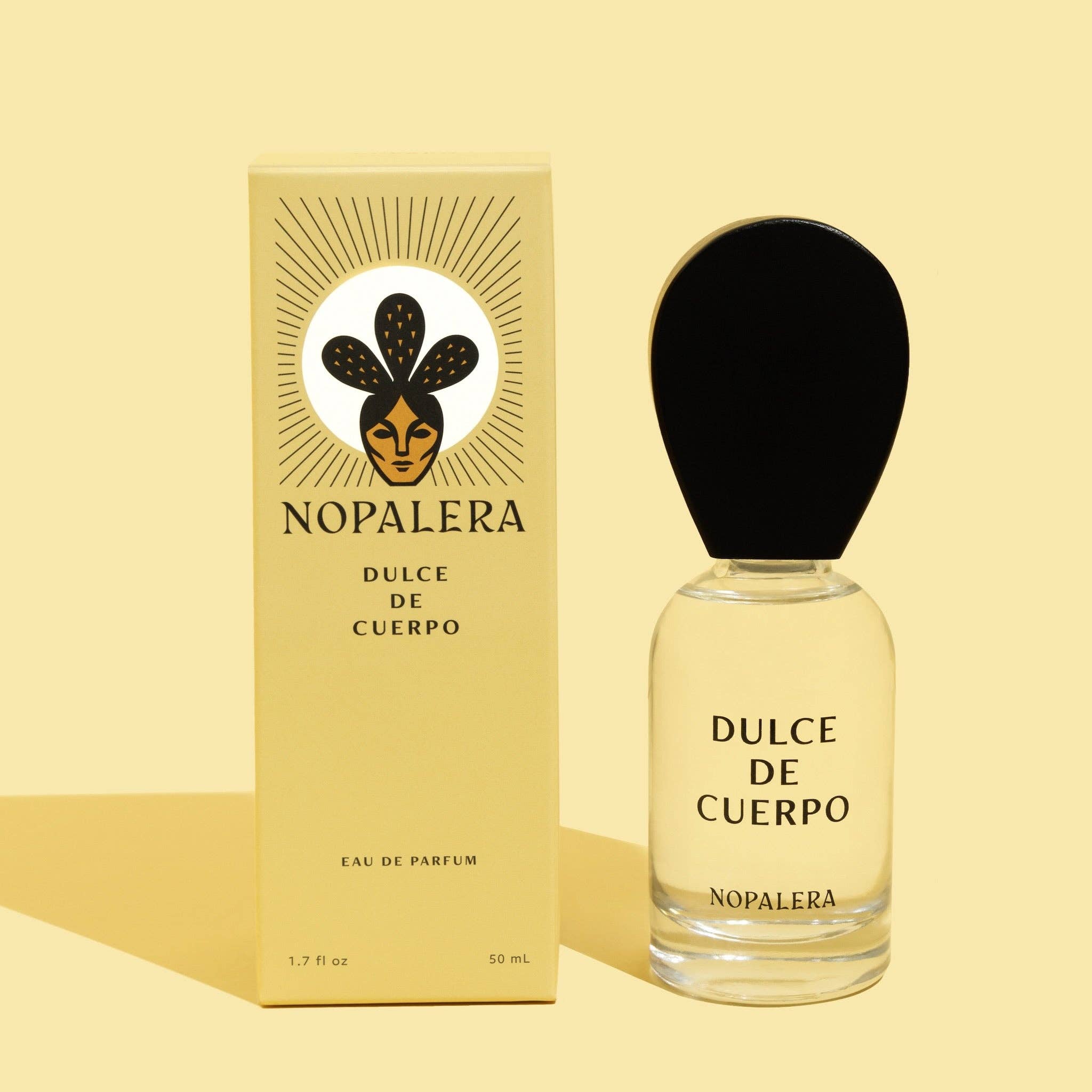 A 50ml bottle of Nopaleras Dulce de Cuerpo perfume, with a large black oval cap, stands by its pale yellow box featuring black text and a stylized face logo.