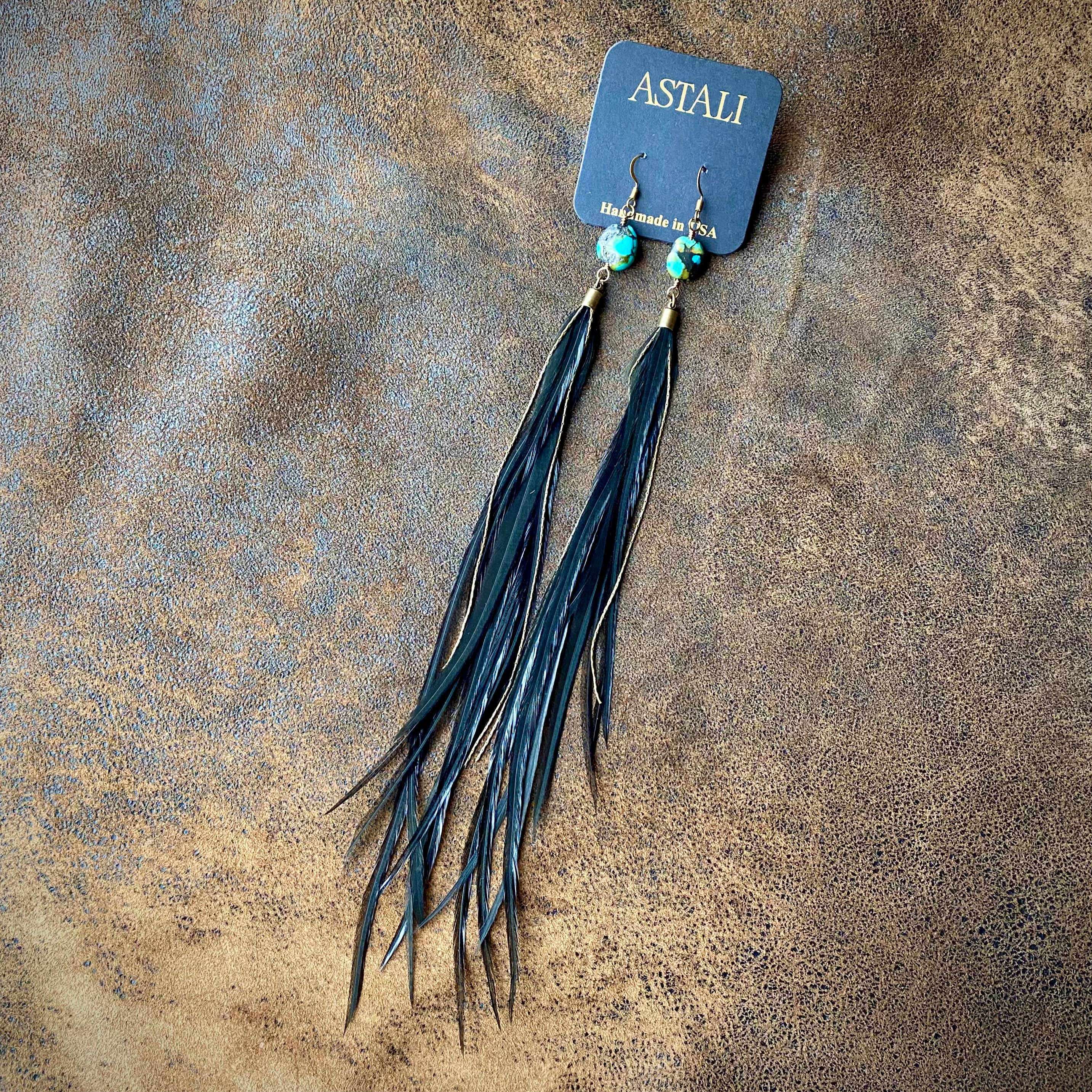 A pair of Feather & Turquoise Statement Earrings by ASTALI, featuring antique brass and sustainable coque feathers, displayed on a card reading Handmade in USA on a textured brown surface.