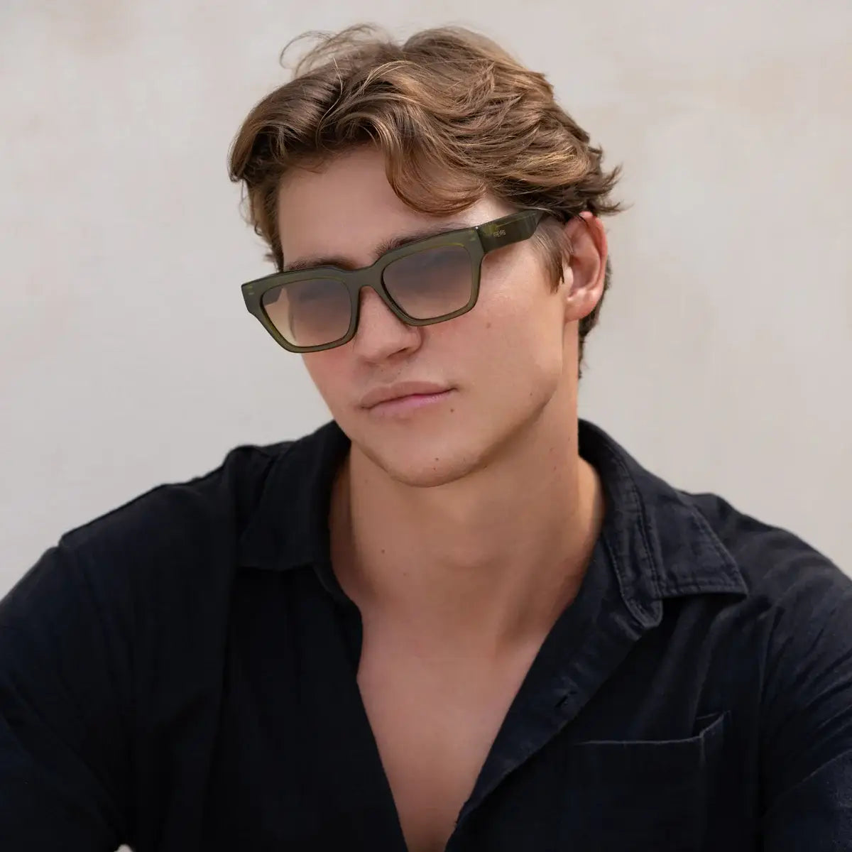 A young man with wavy brown hair wears FREYRS Eyewear Hayden Acetate Mens Rectangular Sunglasses with handmade acetate frames and a black collared shirt, posing against a simple light backdrop.
