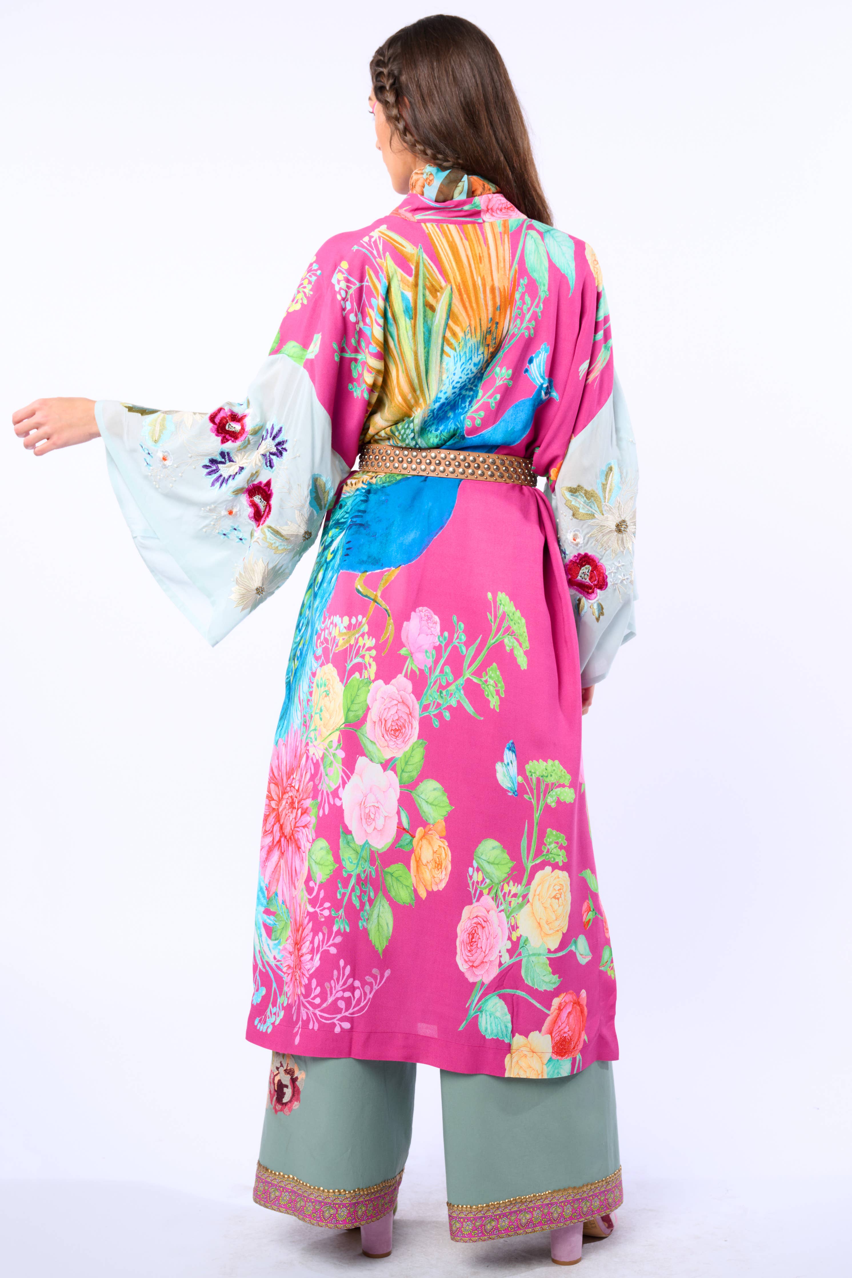 A woman stands facing away, wearing the Aratta Marie Antoinette Kimono with vibrant pink, blue, and green patterns, wide embroidered sleeves, a gold belt, green pants, and pink platform shoes for a bold statement look.