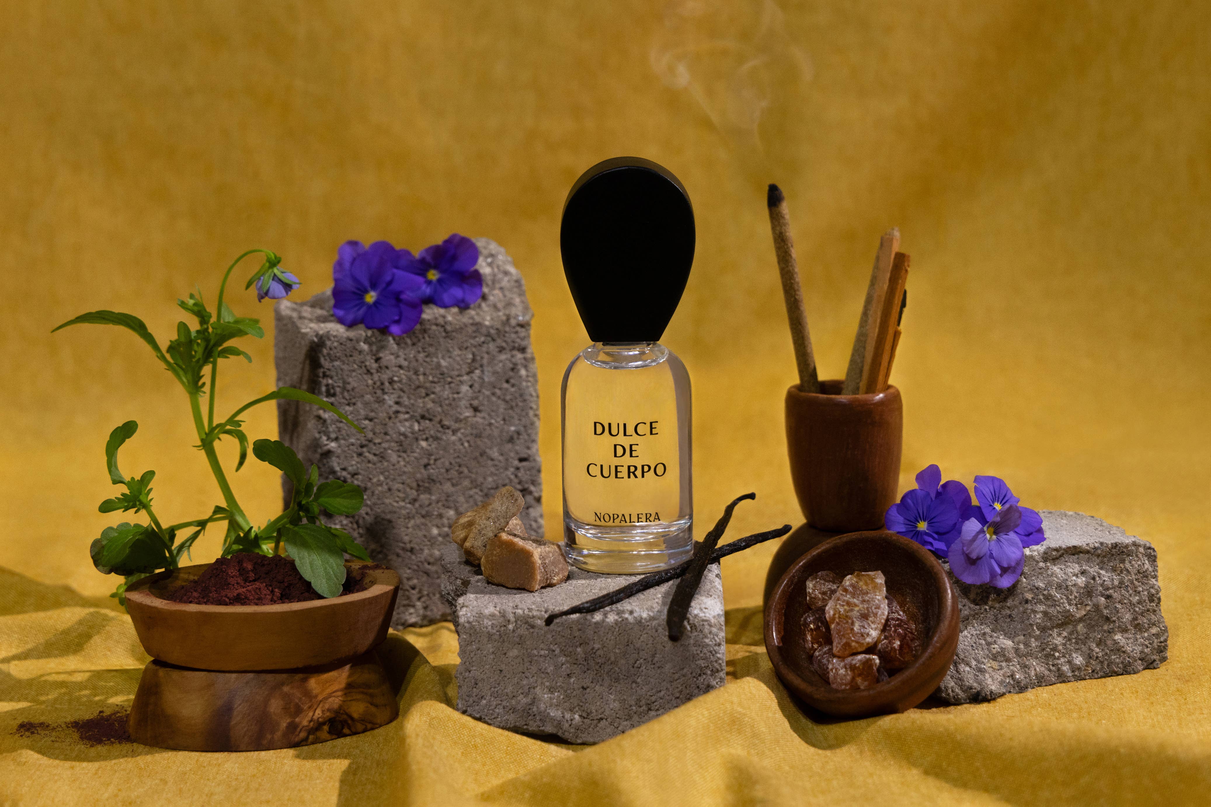 A 50ml Perfume - Dulce de Cuerpo by Nopalera sits on a stone block, surrounded by purple flowers and vanilla beans, showcasing its signature blend of raw brown sugar, cinnamon sticks, and a hint of clean fragrance on a yellow surface.