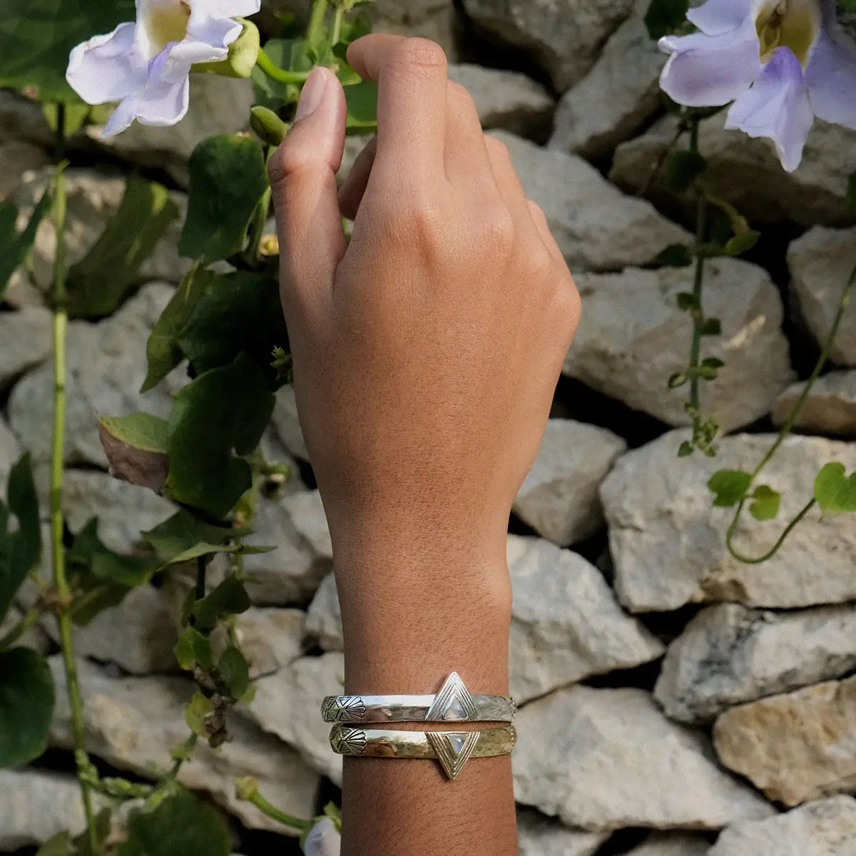 Hand wearing Hold Space Cuff bracelets with geometric design against stone wall and greenery background