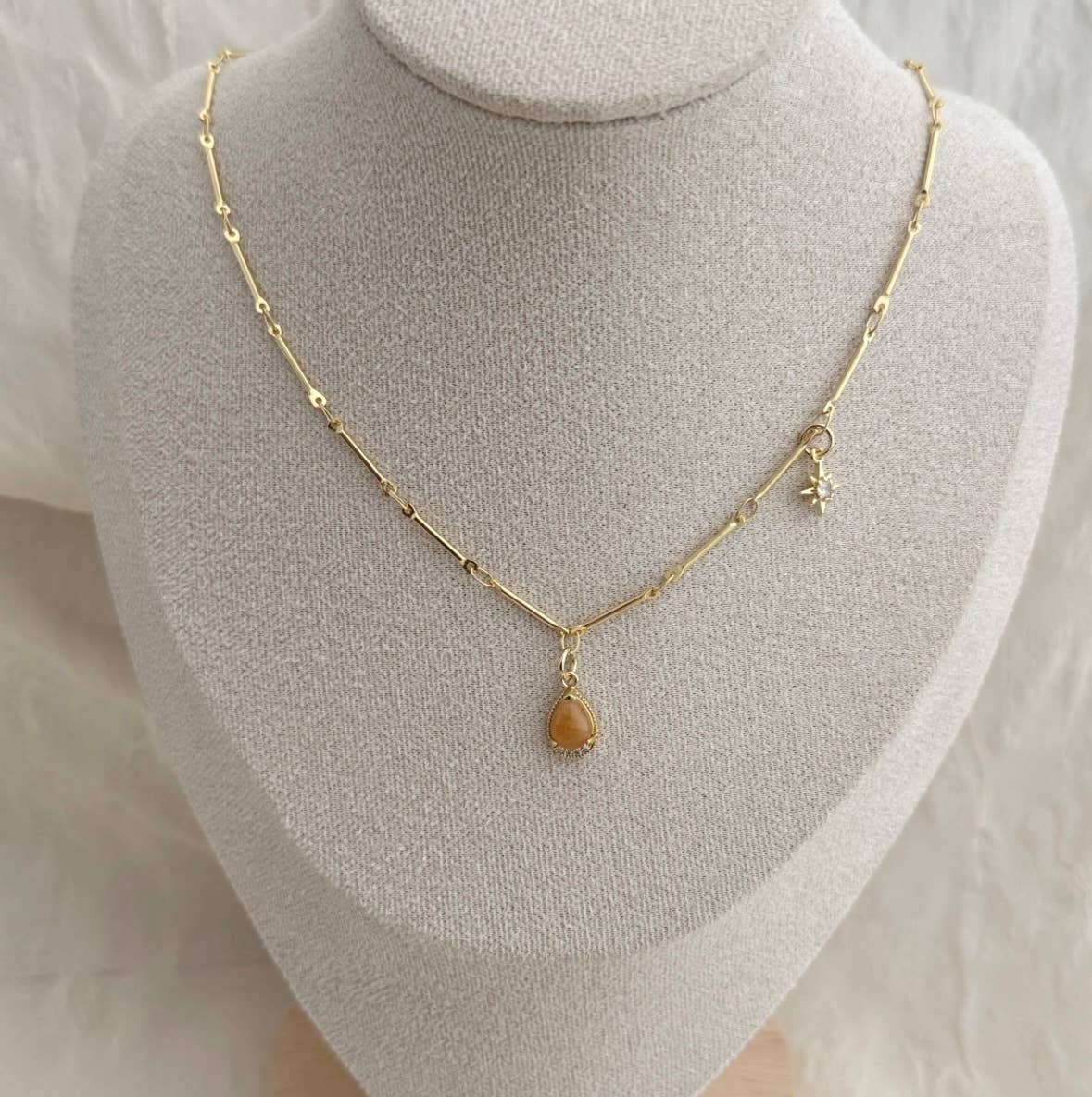 The Stone From The Sun Necklace by Tramps+Thieves features a teardrop amber pendant and star charm on an 18K gold-filled chain, shown on a beige bust.