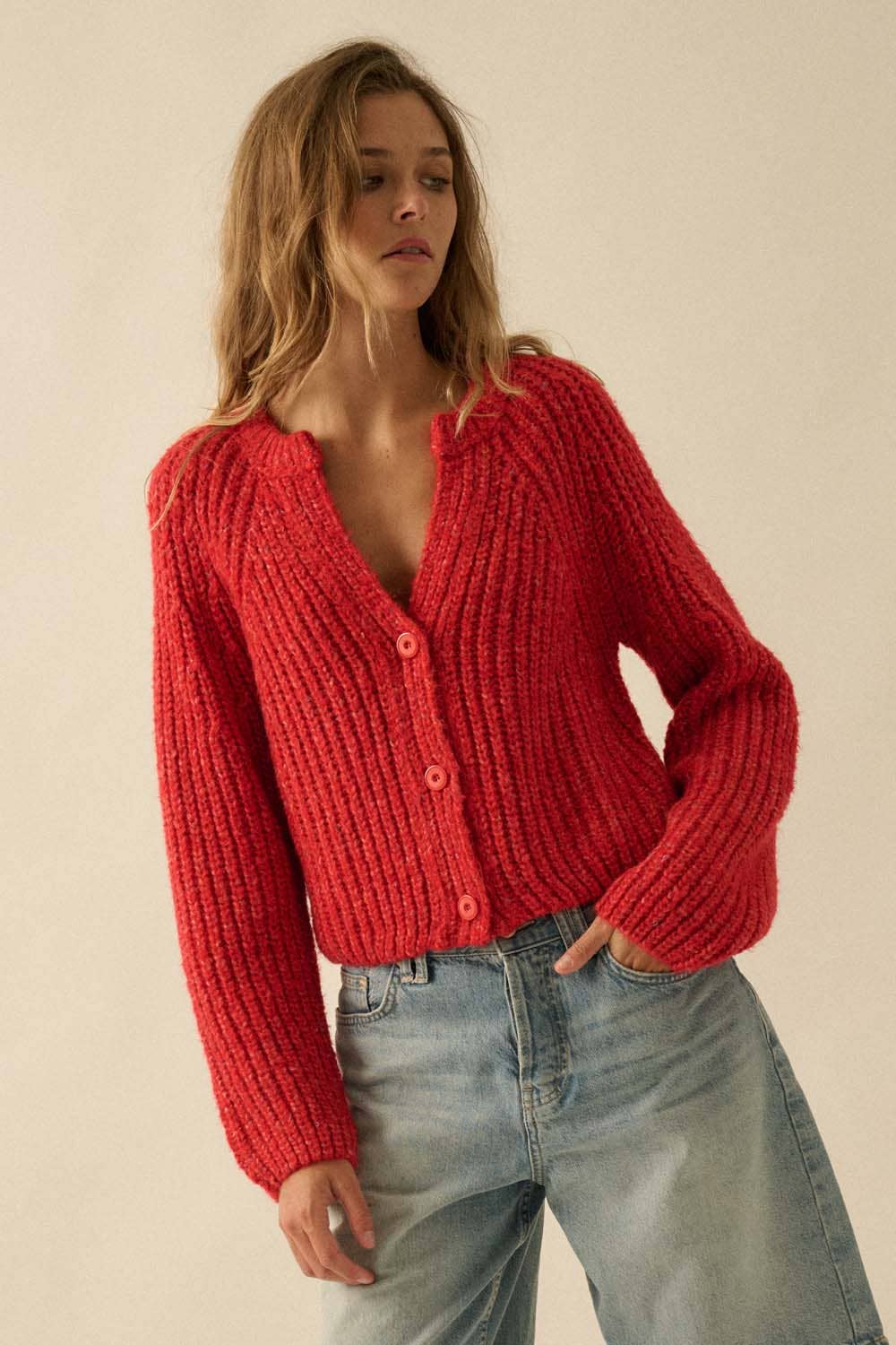 A person wears the Confetti Button-Front Cardigan Sweater by Promesa USA, featuring a chunky red ribbed knit, large sleeves, and blue jeans, standing against a plain beige background.