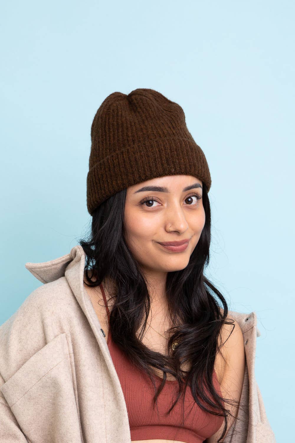 A young woman with long dark hair smiles softly in front of a light blue background, wearing a Leto Accessories Versatile Cuffed Beanie – Cozy Rib Knit Hat, a beige jacket, and a rust-colored top.