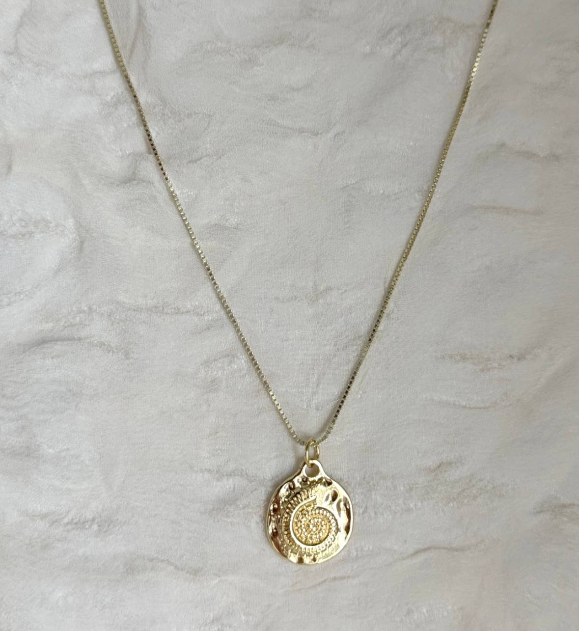 The Mar Medallion Necklace by Tramps+Thieves features a round, textured gold vermeil pendant with an intricate spiral design, shown on a light, textured fabric background.