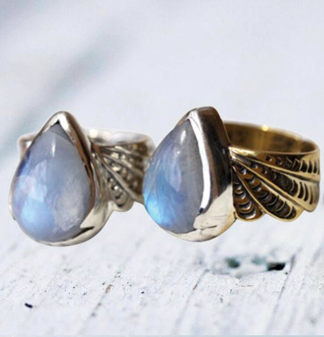 Two bahgsu jewels Winged Rings showcase large, teardrop-shaped moonstones with a blue sheen on textured white wood. Each adjustable brass ring features intricate designs, adding elegant charm to any look.