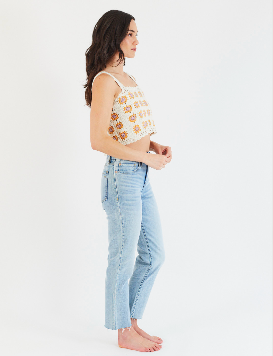A woman with long dark hair stands in profile against a white background, barefoot and relaxed, wearing Porter Blue Apparel’s REBEL STRAIGHT IN EDEN WASH high-rise organic cotton jeans and a sleeveless floral crocheted crop top.