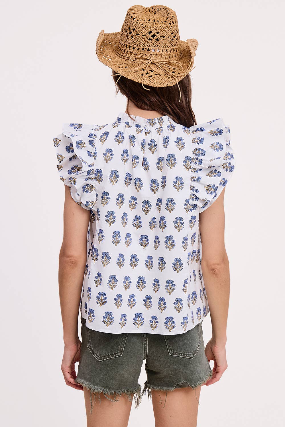 A woman stands with her back to the camera against a light background, wearing a straw hat, green frayed shorts, and the Ultramarine Celeste Flutter Sleeve Blouse in white floral.