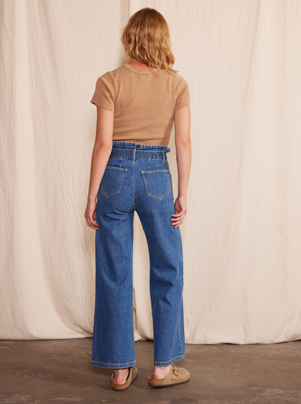 A person with shoulder-length blonde hair, in a beige short-sleeve top and Porter Blue Apparel’s PENNY PAPERBAG WIDE LEG IN LAGUNA WASH jeans, stands facing away against a neutral fabric backdrop, wearing beige sandals.