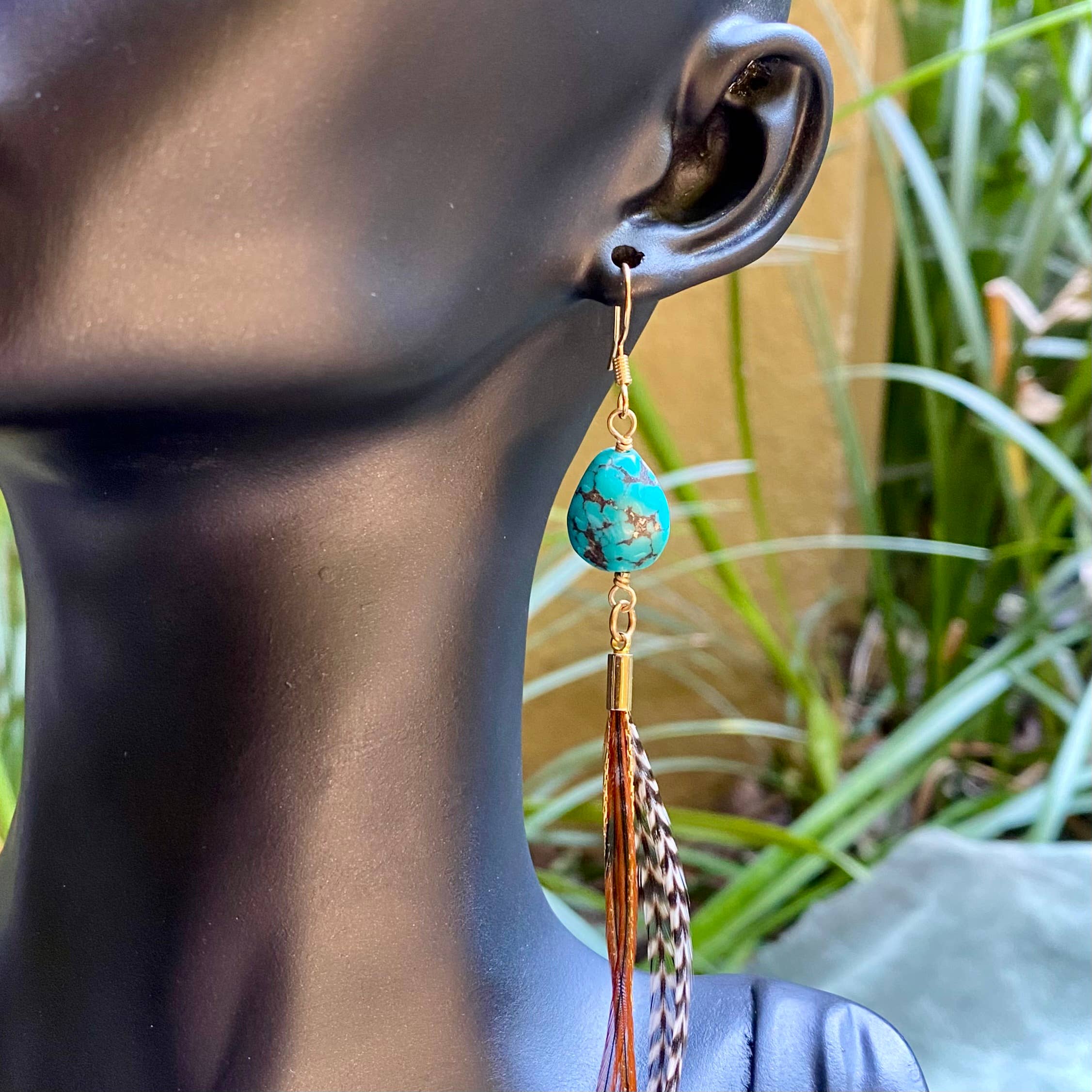 A mannequin head showcases ASTALIs Turquoise & Mini Feather Earrings—Grizzly & Fawn/Gold—with a teardrop turquoise stone, gold-plated hardware, and long multicolored feather tassels, set before a green, blurred backdrop.