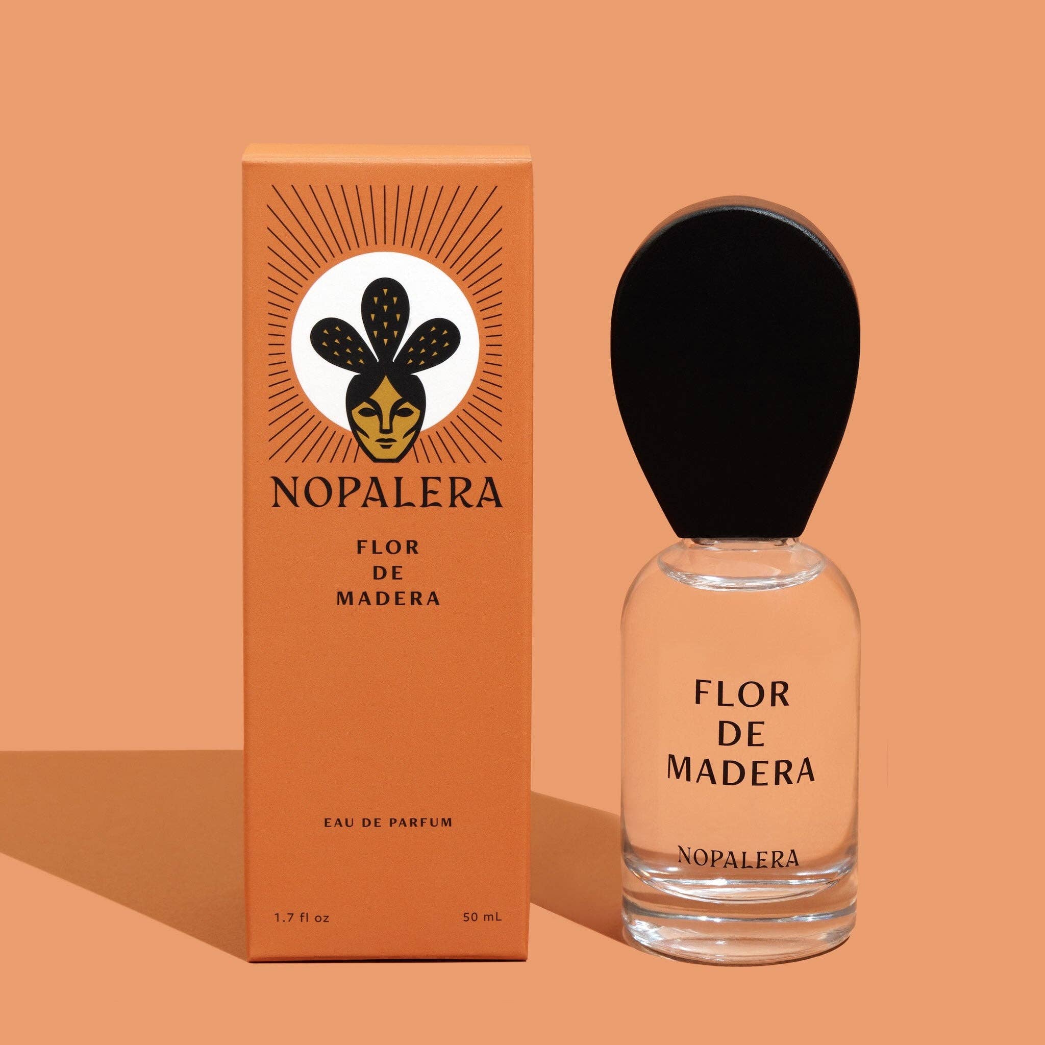 A 10mL Nopalera Flor de Madera Eau de Parfum Travel Spray with Mexican citrus notes stands by its orange box, which features a stylized face and black leaf design. The bottle is topped with a black oval cap.
