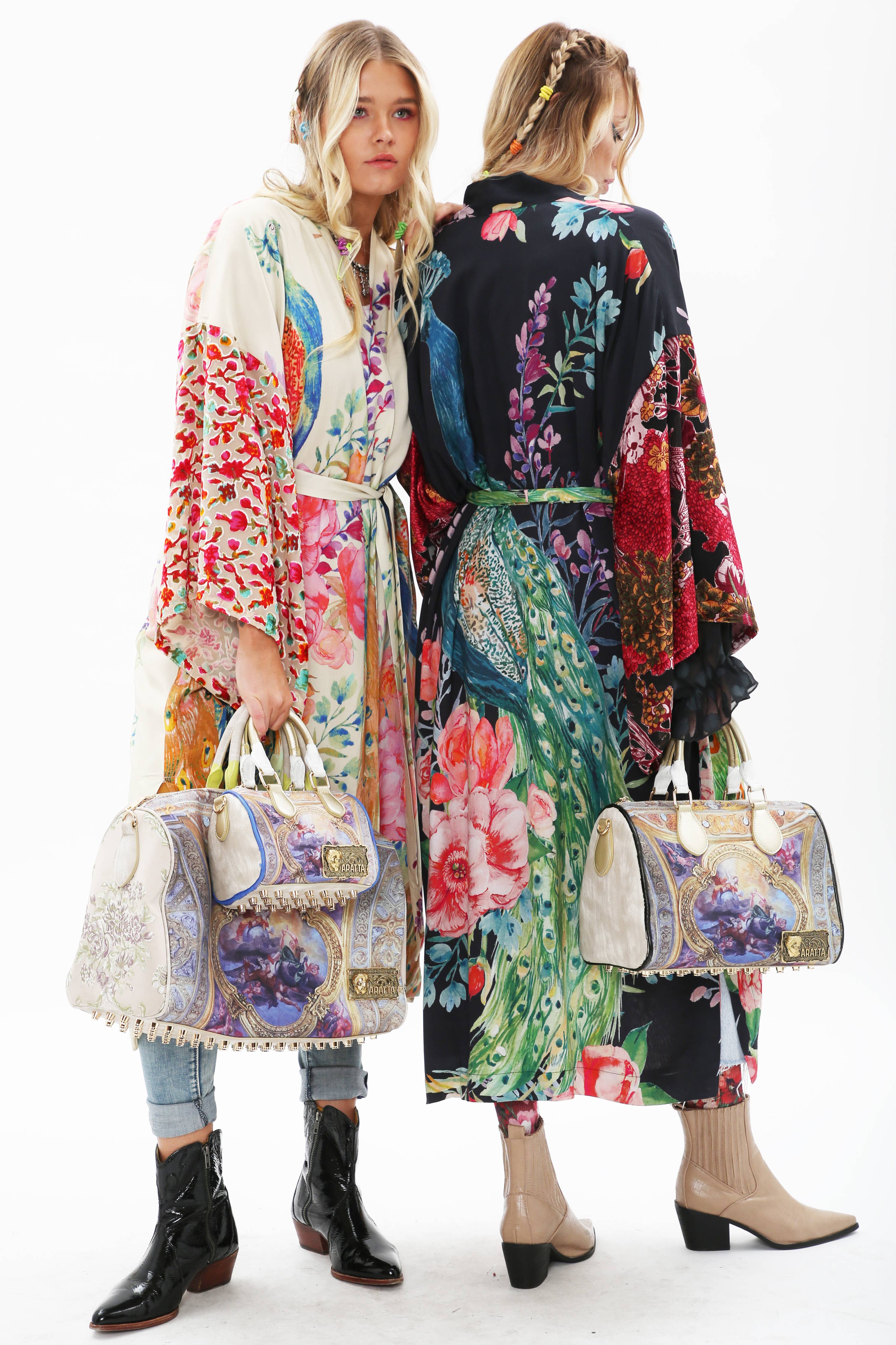 Two women with blonde hair wear Aratta’s Sweet Fantasy Kimono, featuring vibrant peacock floral patterns and burnout velvet sleeves. Each holds a vintage-inspired bag; one faces forward, the other has her back turned. Both sport casual shoes.