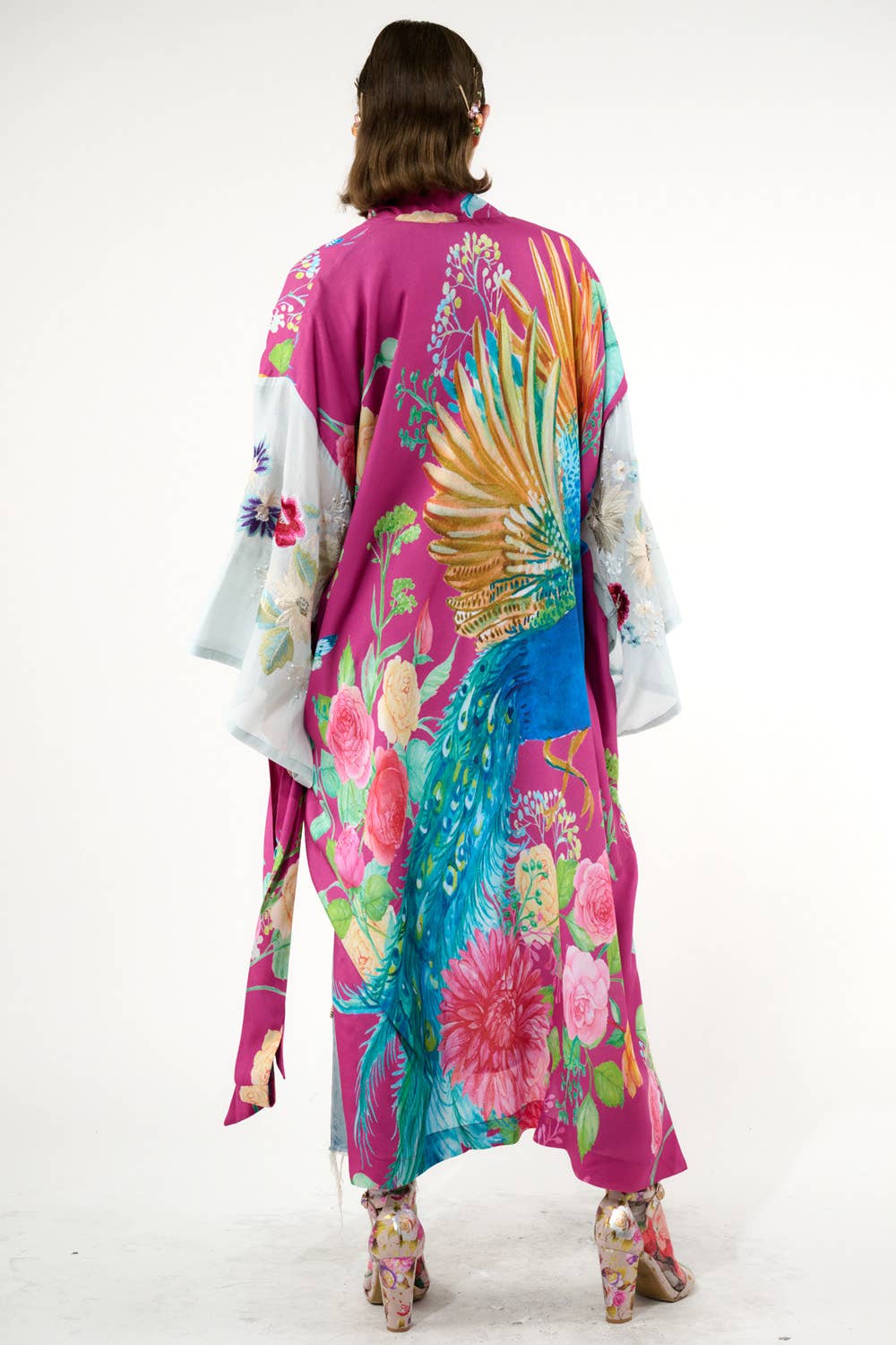 A person stands facing away, wearing the Aratta Marie Antoinette Kimono with embroidered floral sleeves and a vibrant peacock design on the back. They pair it with matching floral heels against a plain white background, highlighting this statement piece.