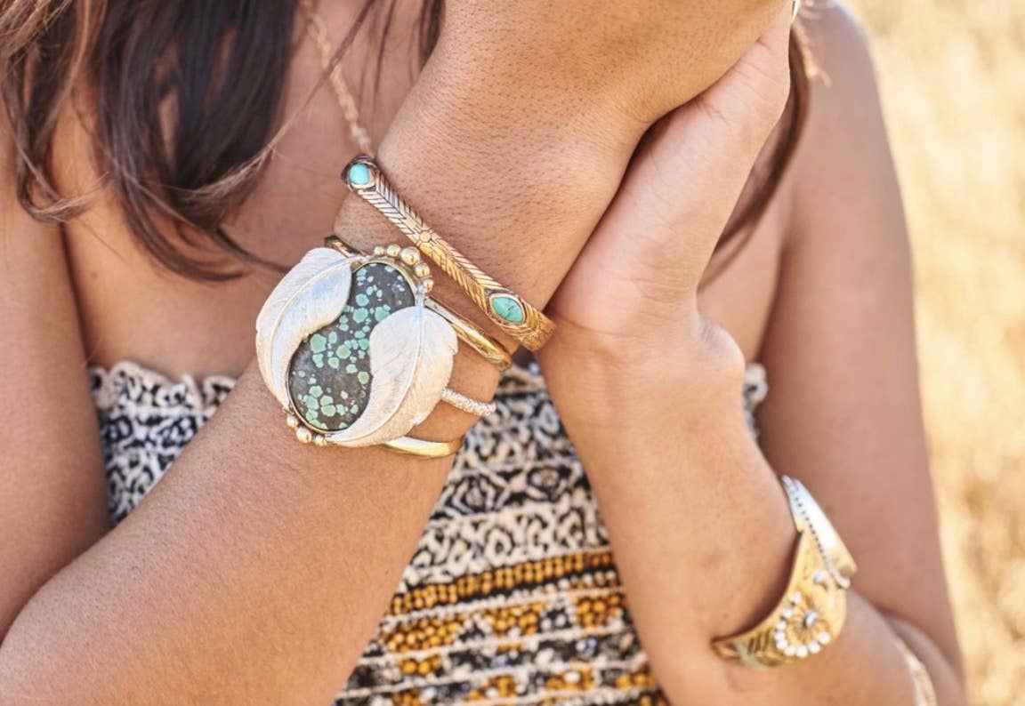 A woman wearing several bracelets, including the bahgsu jewels Medicine Wheel Cuff with a bold turquoise inlay, holds her hands near her face. She is dressed in a patterned, strapless top.