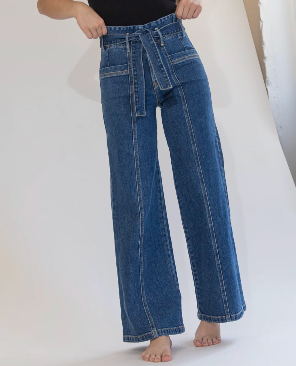 A person stands barefoot on a light background wearing Porter Blue Apparel’s PENNY PAPERBAG WIDE LEG IN LAGUNA WASH—high-waisted, wide-leg blue jeans in organic cotton denim with a matching belt. The upper body isn’t visible.
