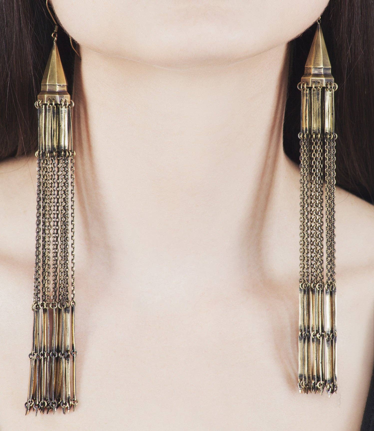 A close-up shows a person with fair skin and dark hair wearing striking, handmade Chandelier Earrings by Jan Hilmer + Sparrow—long dangle brass earrings with multiple gold chains and cylindrical segments extending past the shoulders.