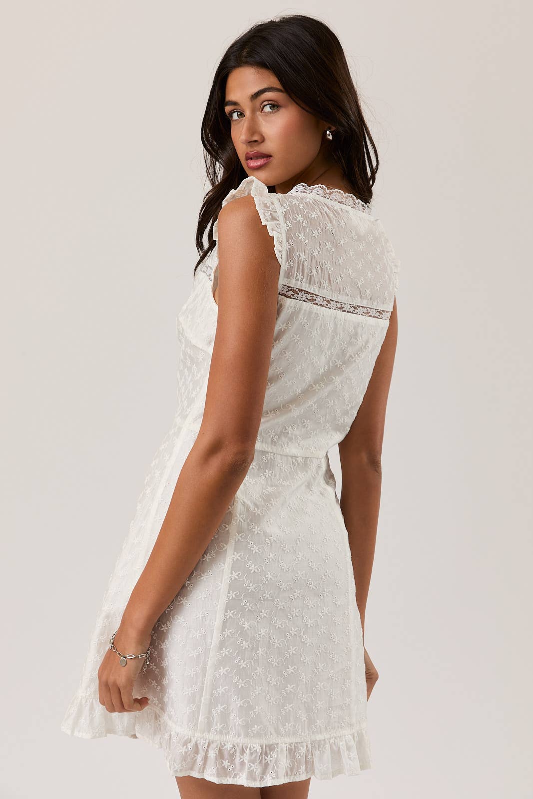 Woman wearing white eyelet lace mini dress with ruffled edges, showcasing back and side details, looking over shoulder
