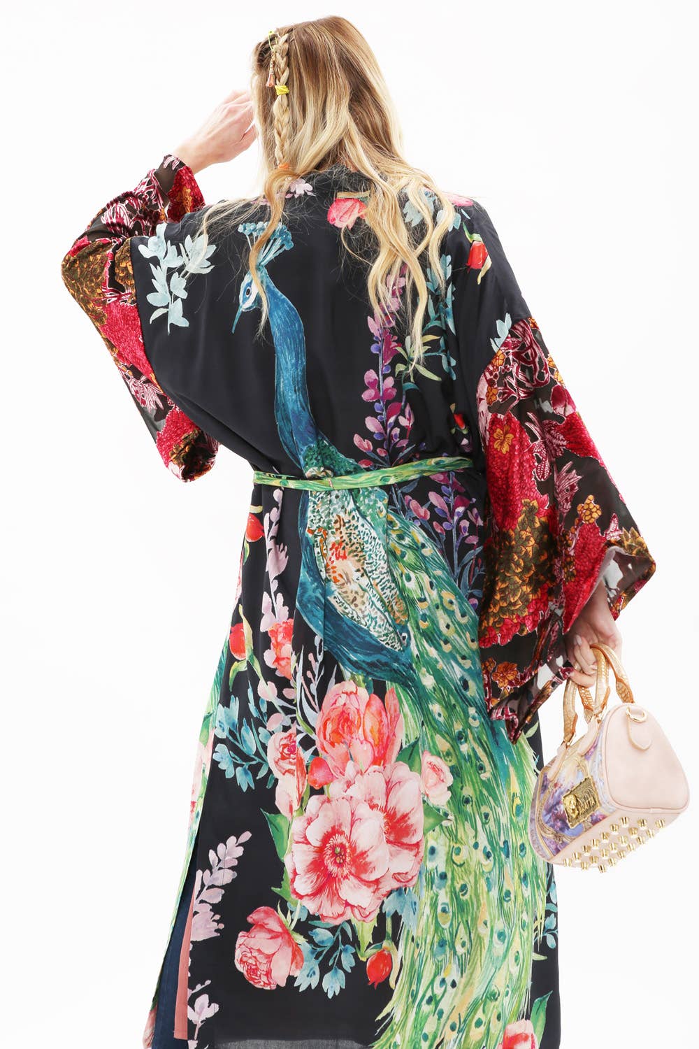 A person with long blonde hair adorned with clips wears the Aratta Sweet Fantasy Kimono, featuring burnout velvet sleeves, and holds a light pink handbag while facing away from the camera against a white background.