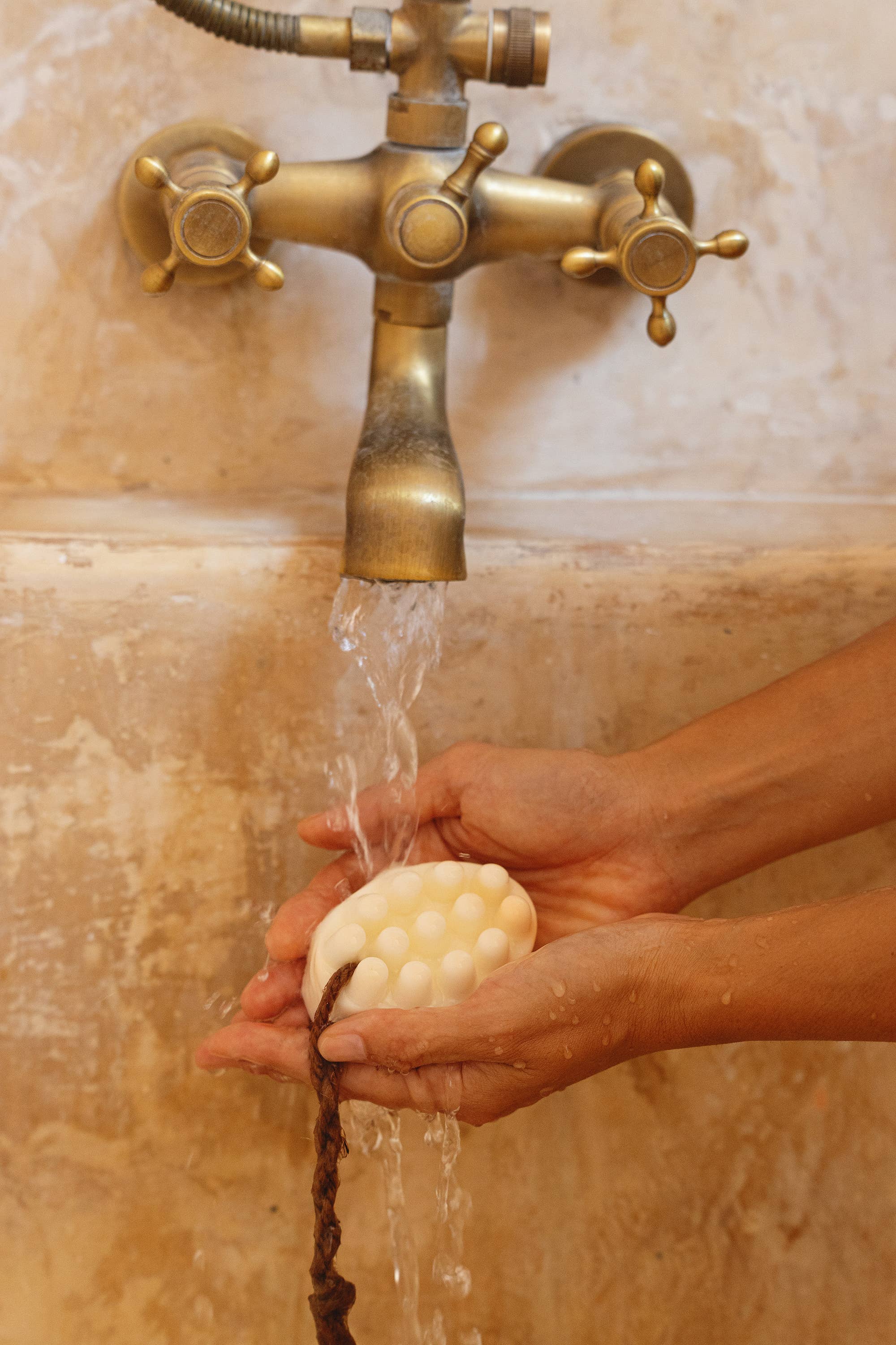 A person holds a Cedar and Myrrh Palo Santo + Shea Butter Bar Soap under running water from a brass faucet over a beige stone sink.
