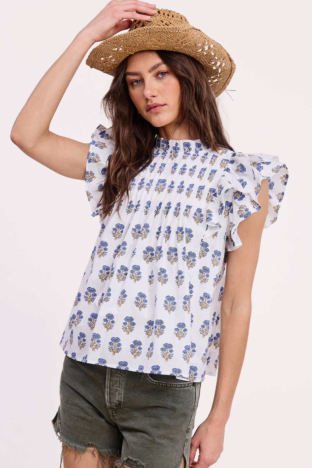 A woman wears the Ultramarine Celeste Flutter Sleeve Blouse with a blue and yellow floral print, olive green frayed shorts, and a straw hat. She holds her hat and looks at the camera against a plain background.
