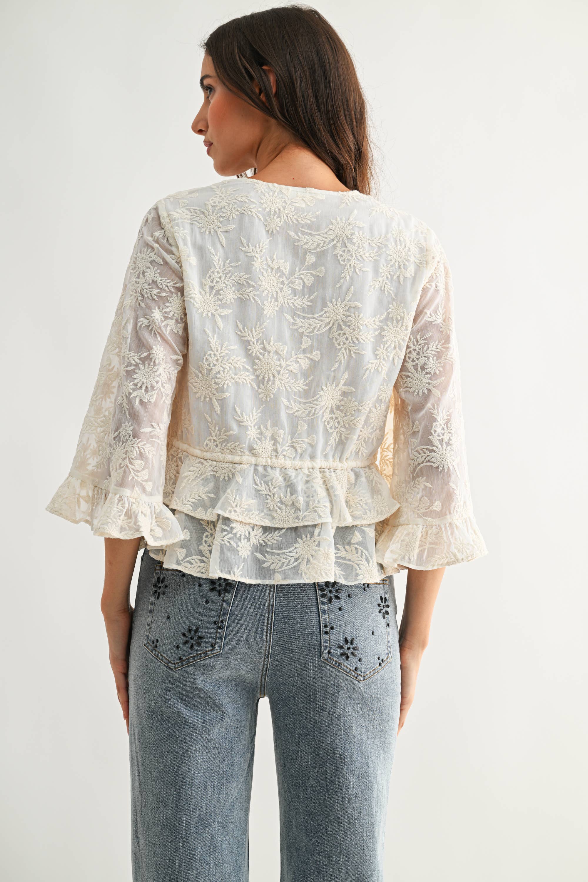 Back view of a woman wearing a cream Boho Embroidered Sheer Blouse with layered ruffle hem and floral embroidery.