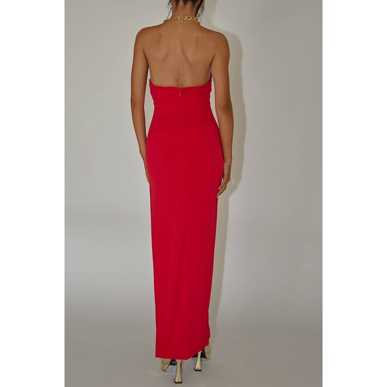 A woman models the One and Only Collective Halterneck Gold Chain Red Maxi Dress, facing away from the camera against a neutral backdrop. She pairs the evening maxi dress with a gold necklace and high heels.