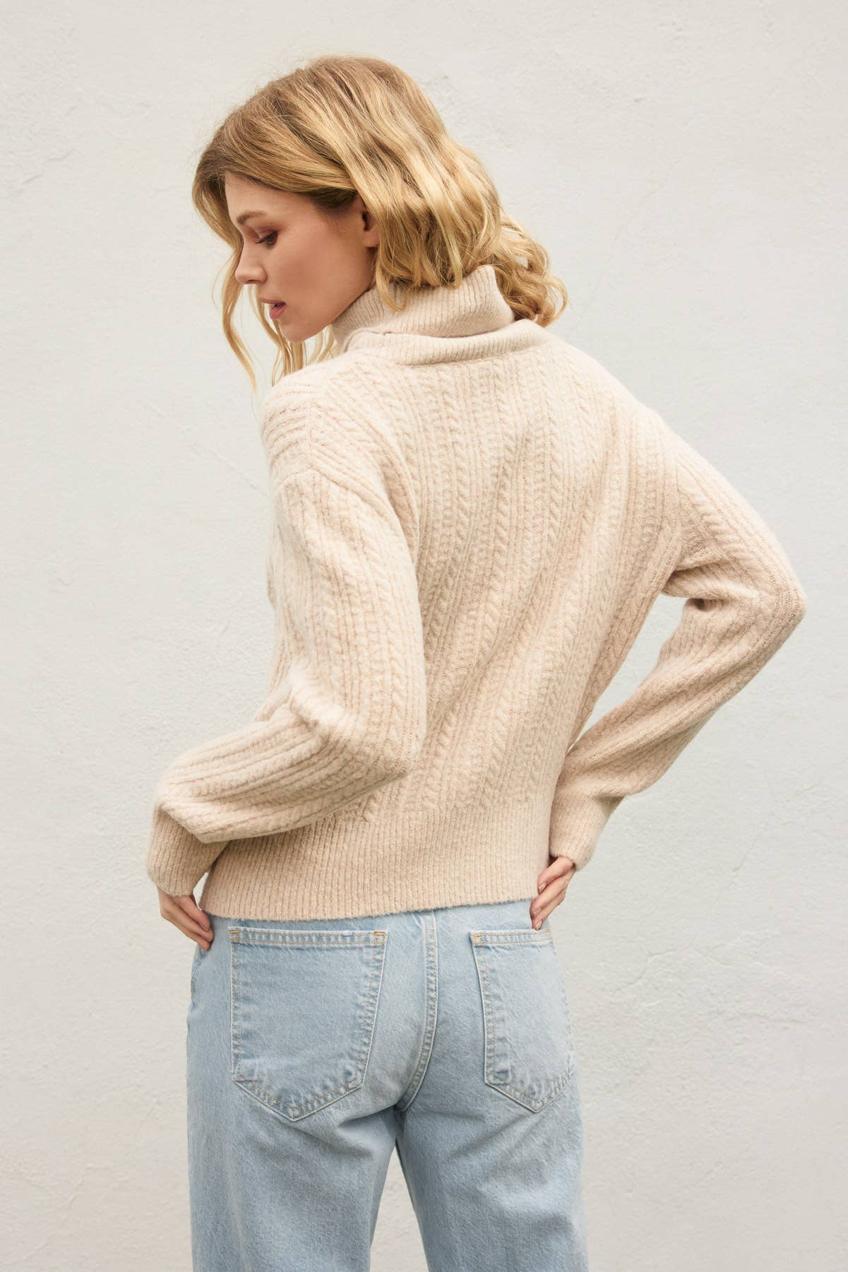 A woman with wavy blonde hair wears the Dress Forum Fluffy Cable Detail Turtle Neck Sweater in beige and light blue jeans, standing with her back to the camera against a plain light background.