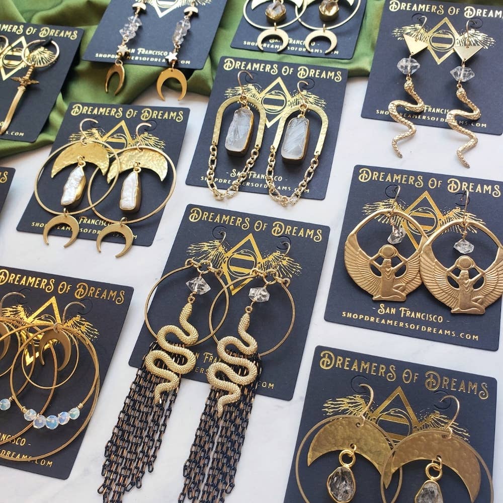 A display of Dreamers of Dreams hypoallergenic earrings—including Rising Snake Earrings, crescent moons, wings, crystals, and beadwork—some adorned with Herkimer diamonds, all arranged on branded cards.