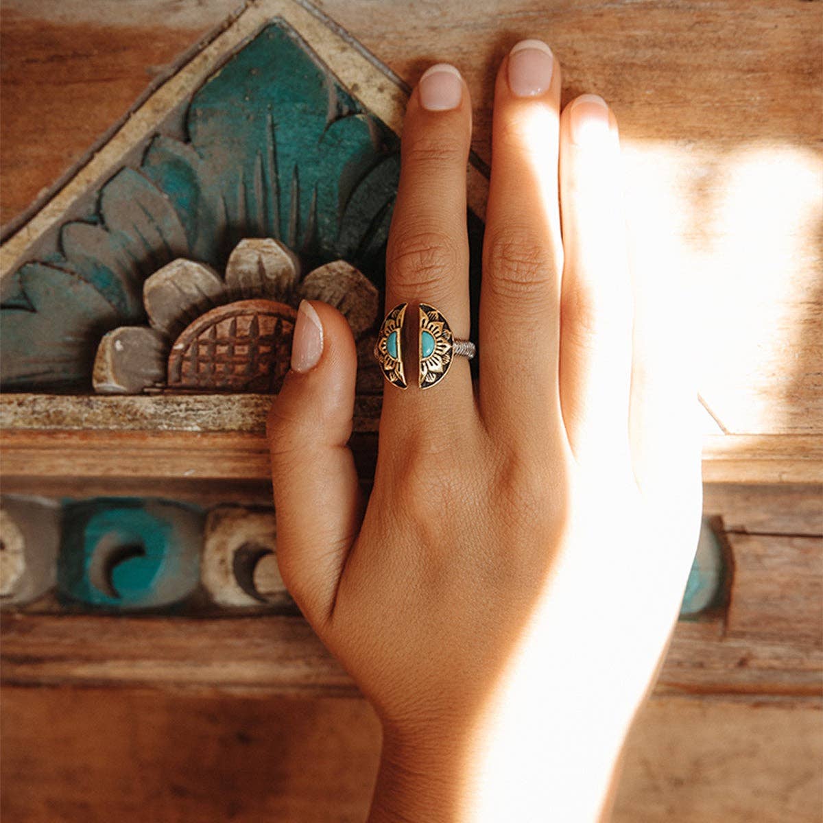A hand with neatly manicured nails wears the bahgsu jewels Natural Mystic Ring - Turquoise, an ornate adjustable piece with intricate designs and a central turquoise stone, as sunlight warms the wooden surface beneath.