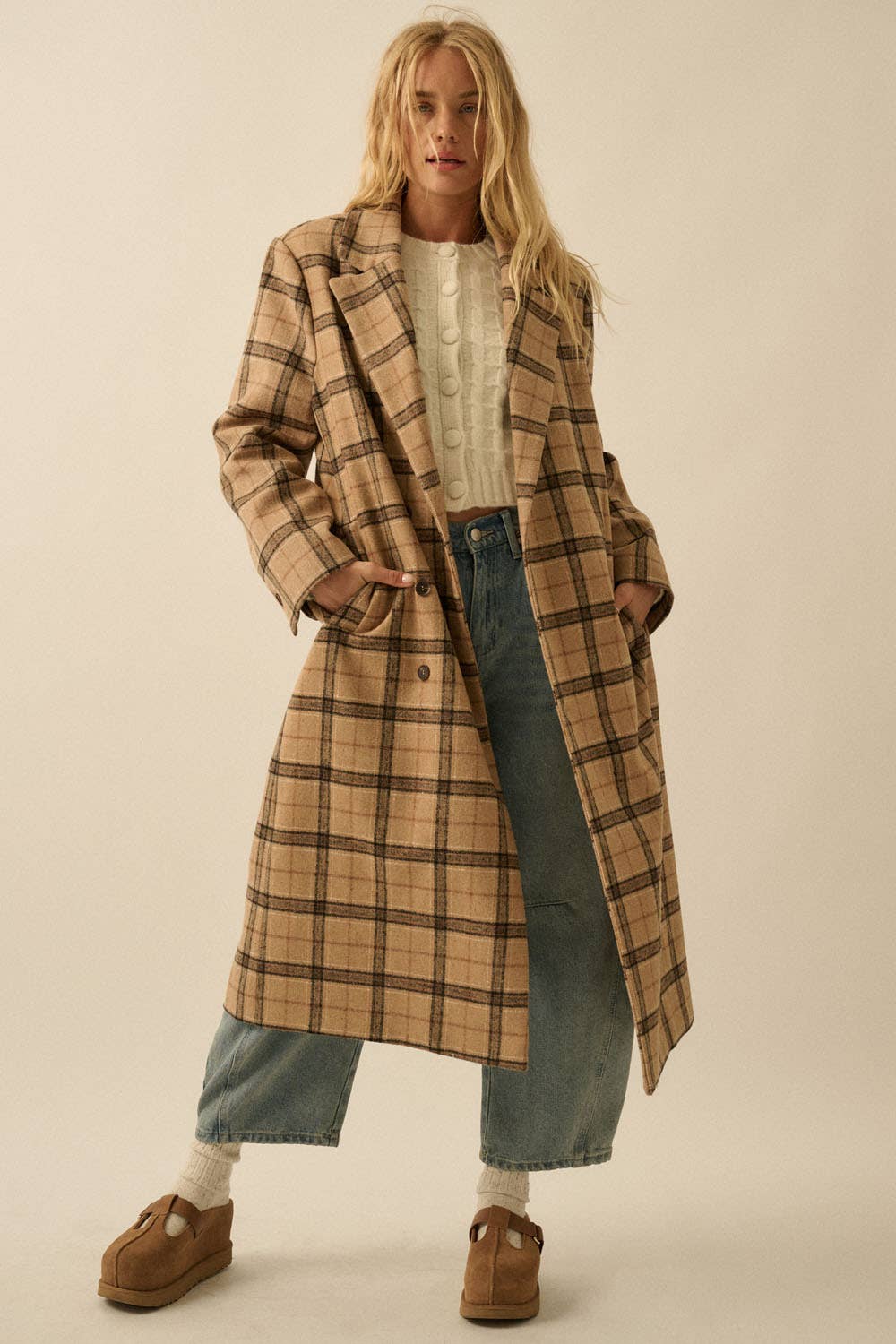 A woman stands facing forward with her hands in her pockets, wearing the Promesa USA Plaid Felt Double-Breasted Calf-Length Overcoat over a white sweater, blue jeans, and brown shoes. Her long blonde hair is loose.