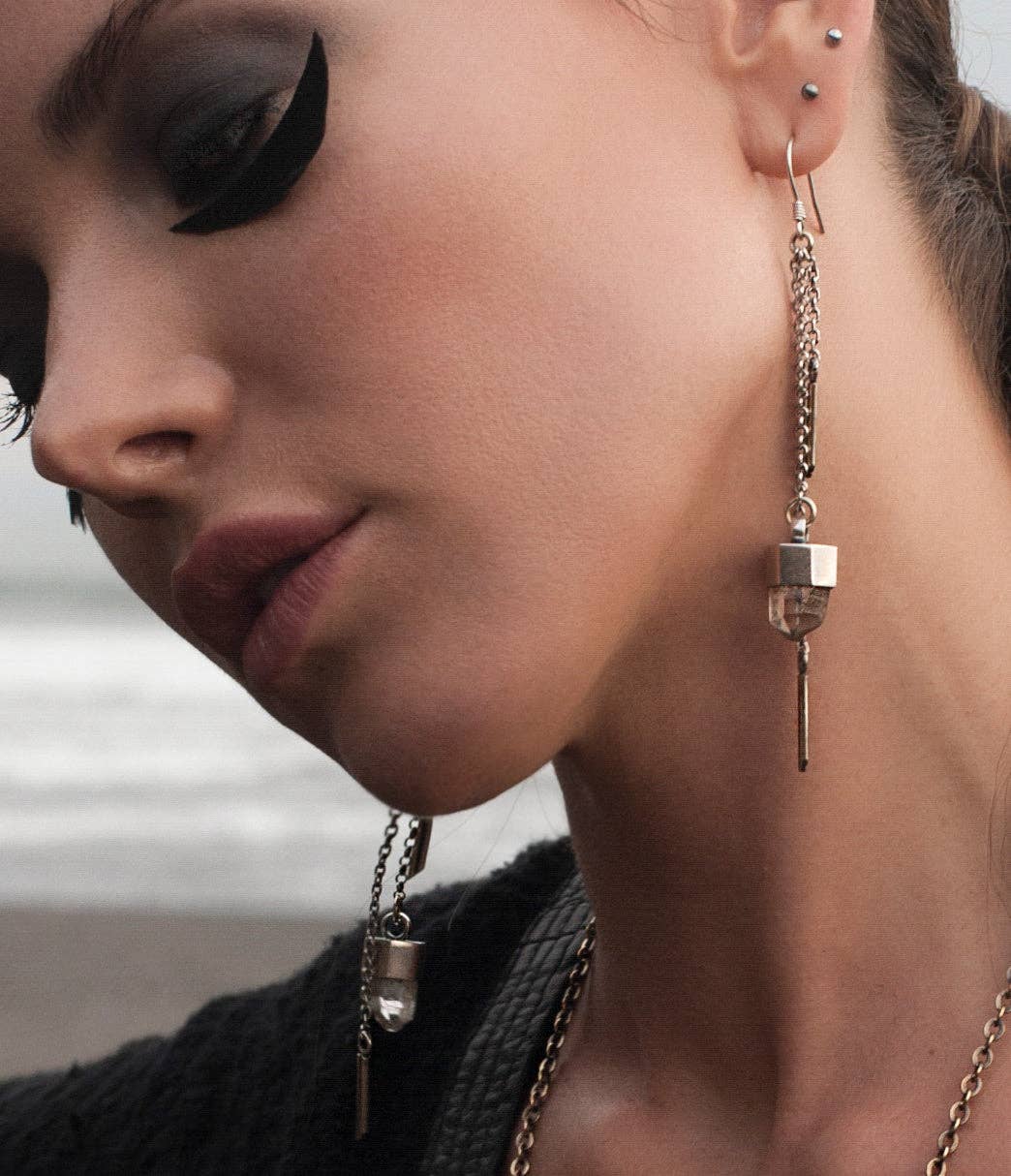 A close-up of a woman with bold black winged eye makeup, wearing Jan Hilmer + Sparrow Crystal dangle earrings in sterling silver, with multiple ear piercings. A blurred beach and ocean serve as the background.