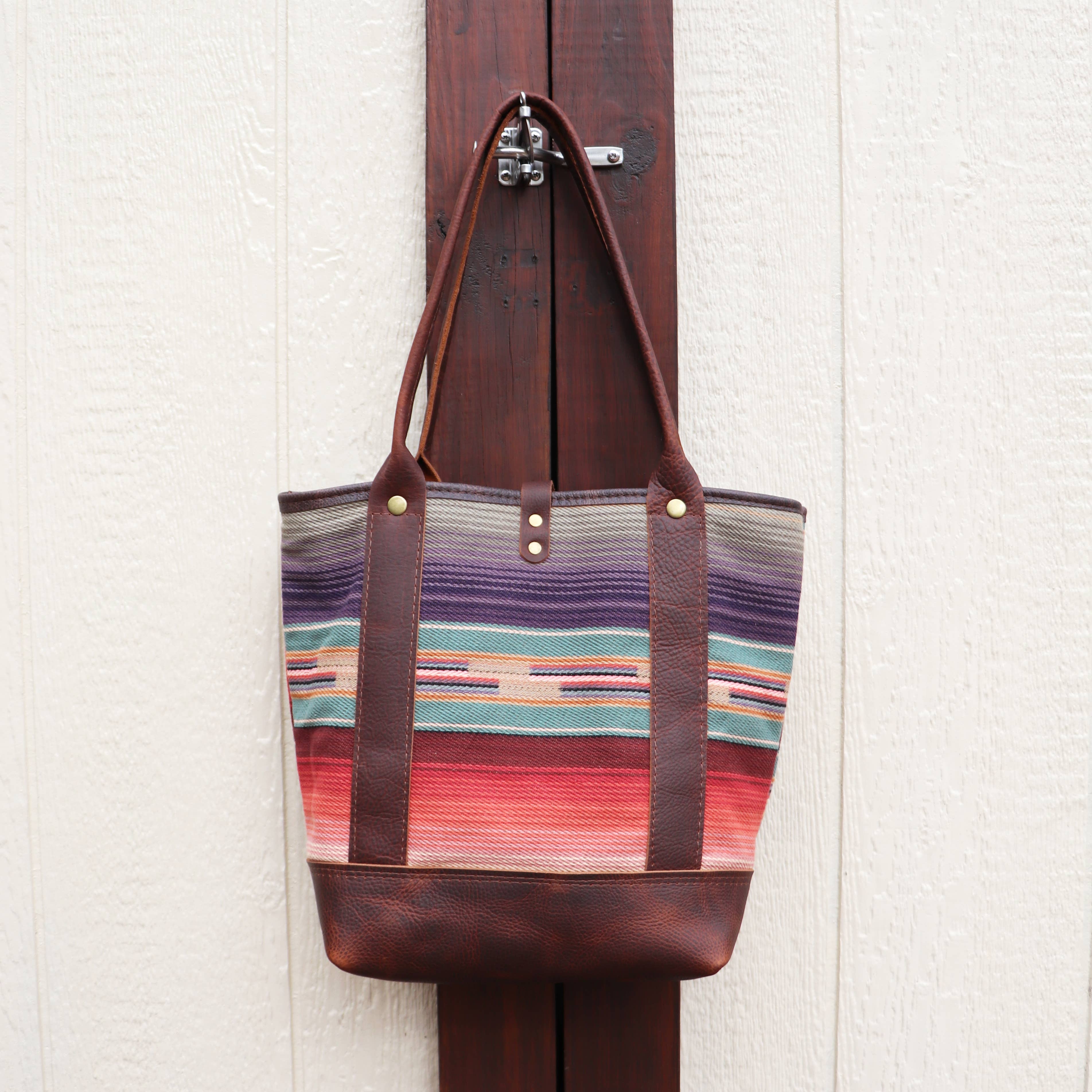 The Pearl Serape Tote by Hawks & Doves, featuring vibrant southwestern serape fabric and brown full grain leather straps and base, hangs on a wooden latch against a white textured wall.