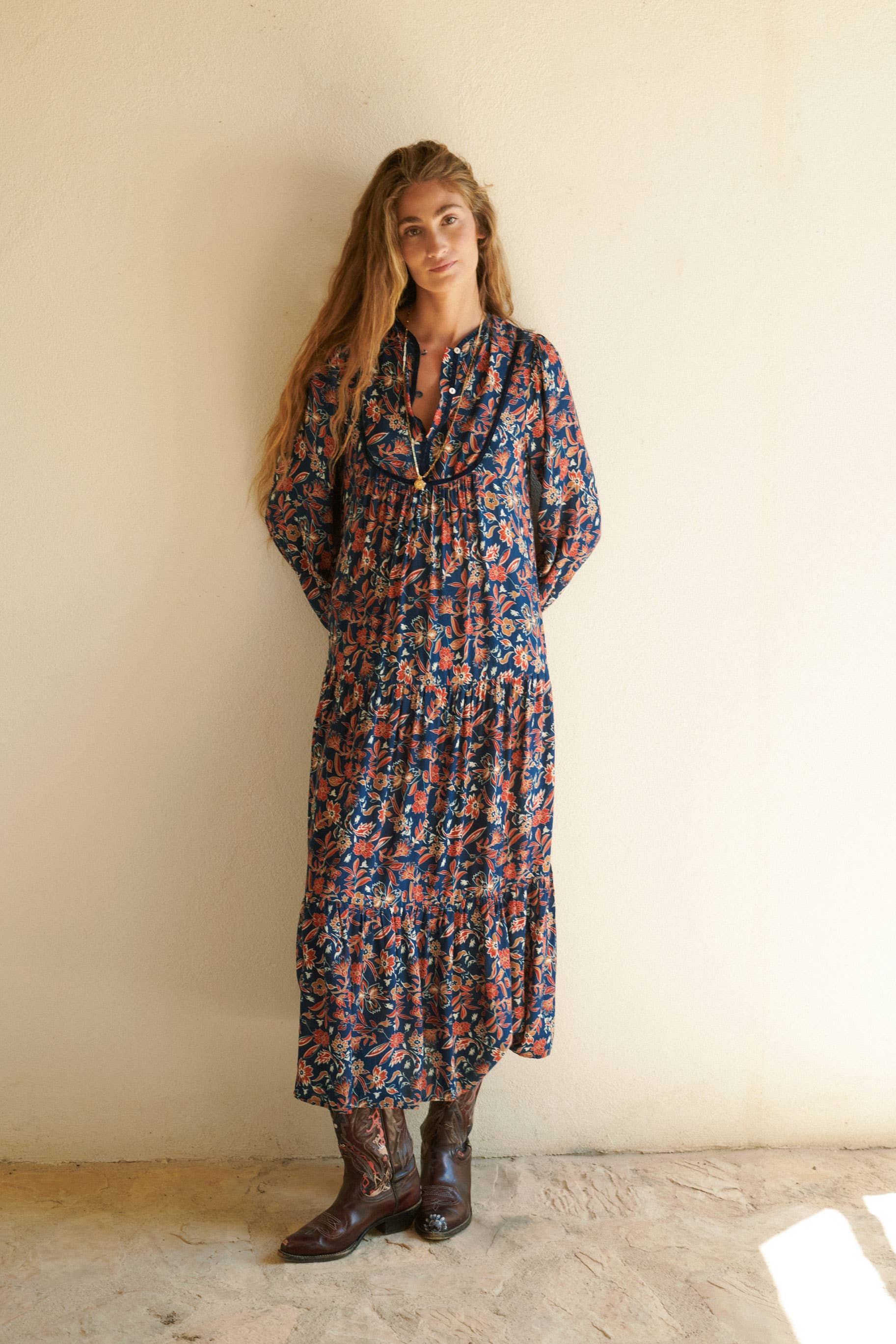 A woman with long, wavy hair stands by a light wall, wearing the Aura Dress Navy by Sonmer Shop USA and brown cowboy boots. She looks relaxed with her hands in her pockets as the bohemian maxi dress flows elegantly around her.