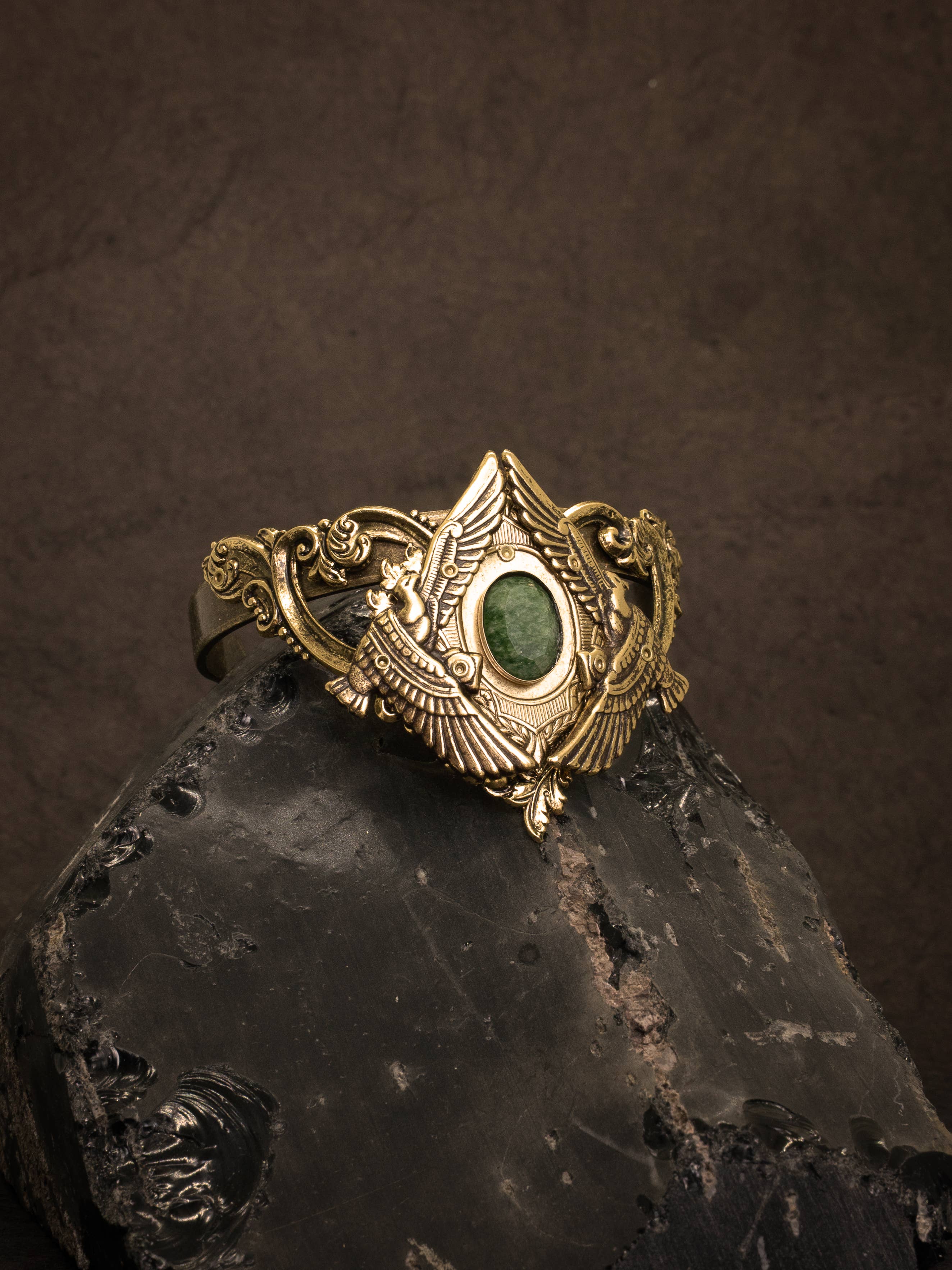 Nekhbet Shield Cuff featuring intricate brass design with central green stone on a dark textured background