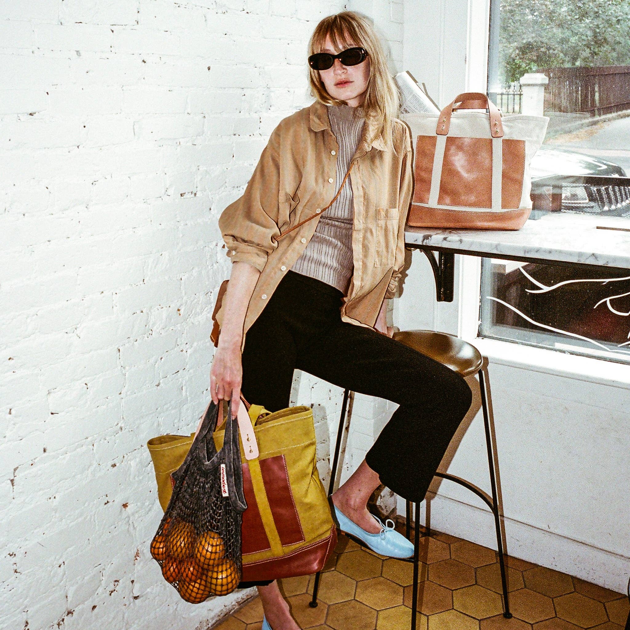 Woman wearing sunglasses sitting indoors with two bags, one clay toned, the other Editor Tote | Chartreuse + Bordeaux