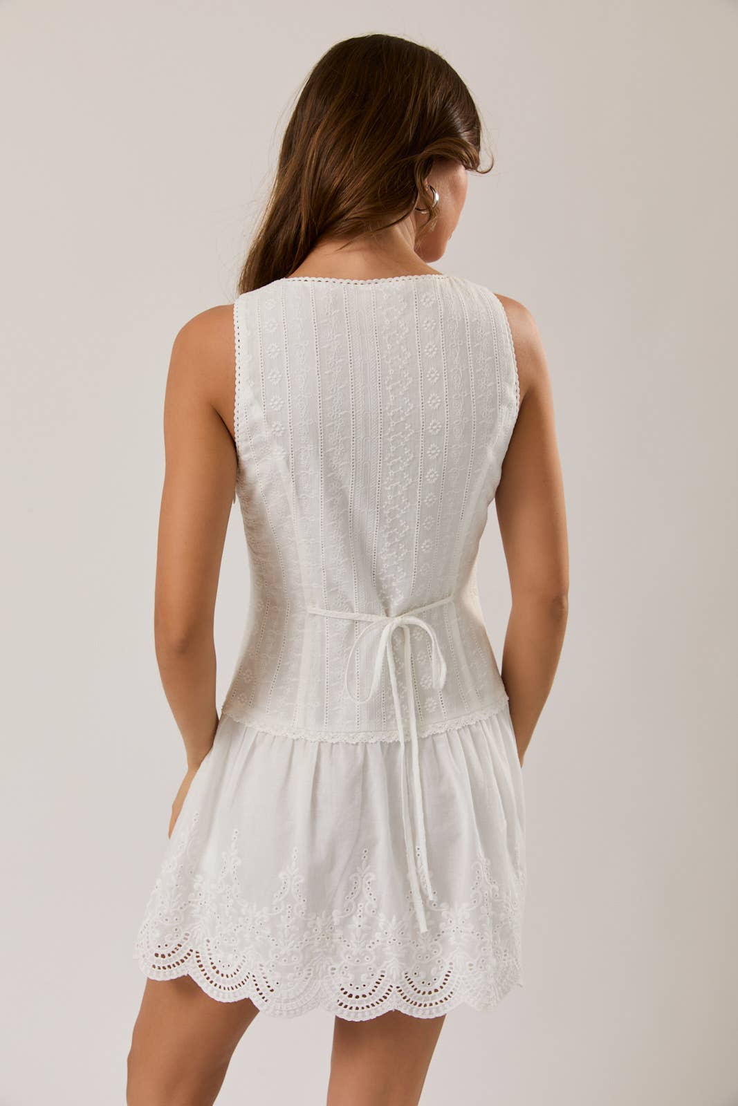 Woman wearing a white sleeveless eyelet mini dress with intricate embroidery and scalloped hem standing with back visible