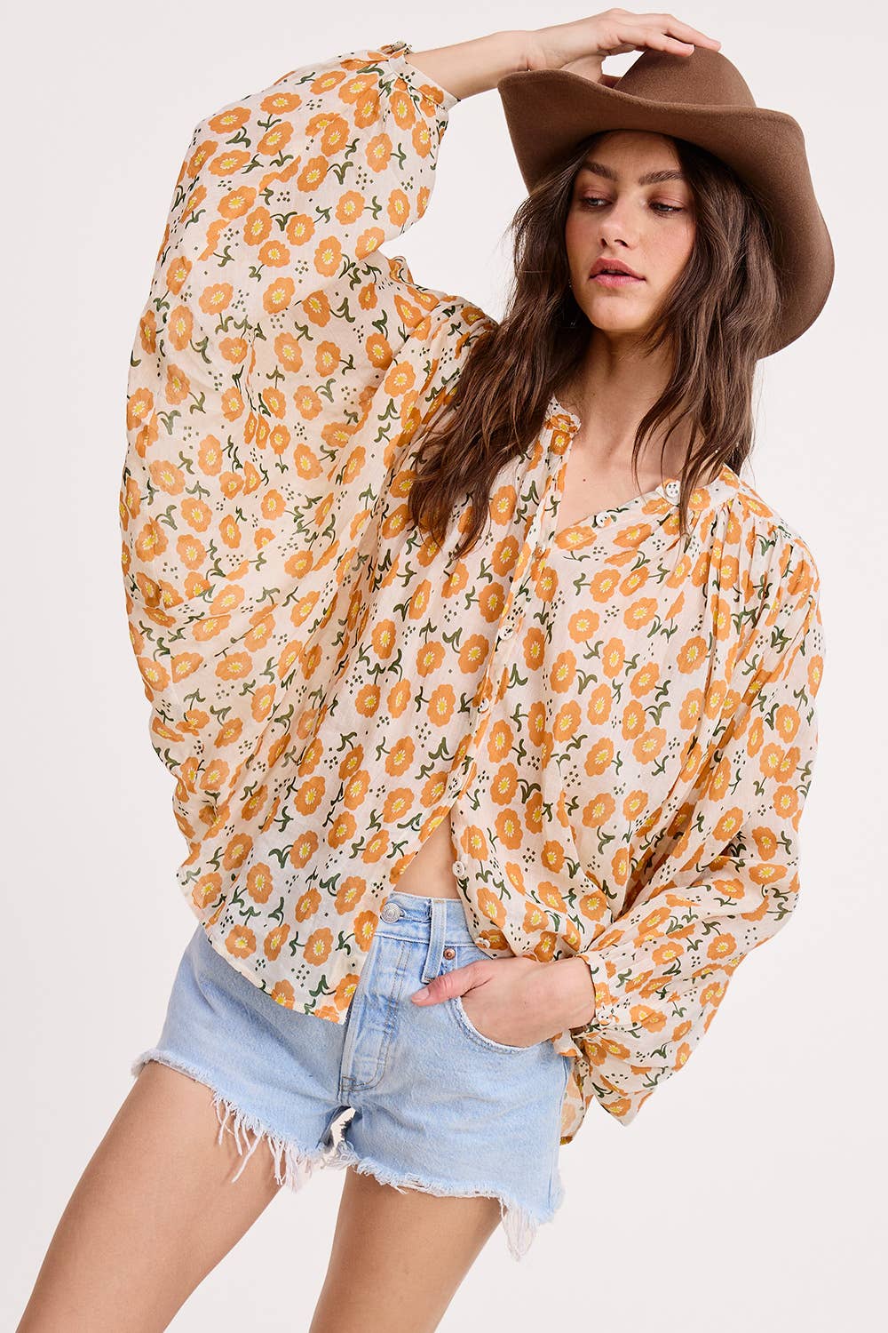 A woman wearing the Ultramarine Meadow Breeze Linen Blouse, featuring orange-yellow floral prints, paired with a brown hat and light blue denim shorts, poses with one hand on her hat against a plain background.