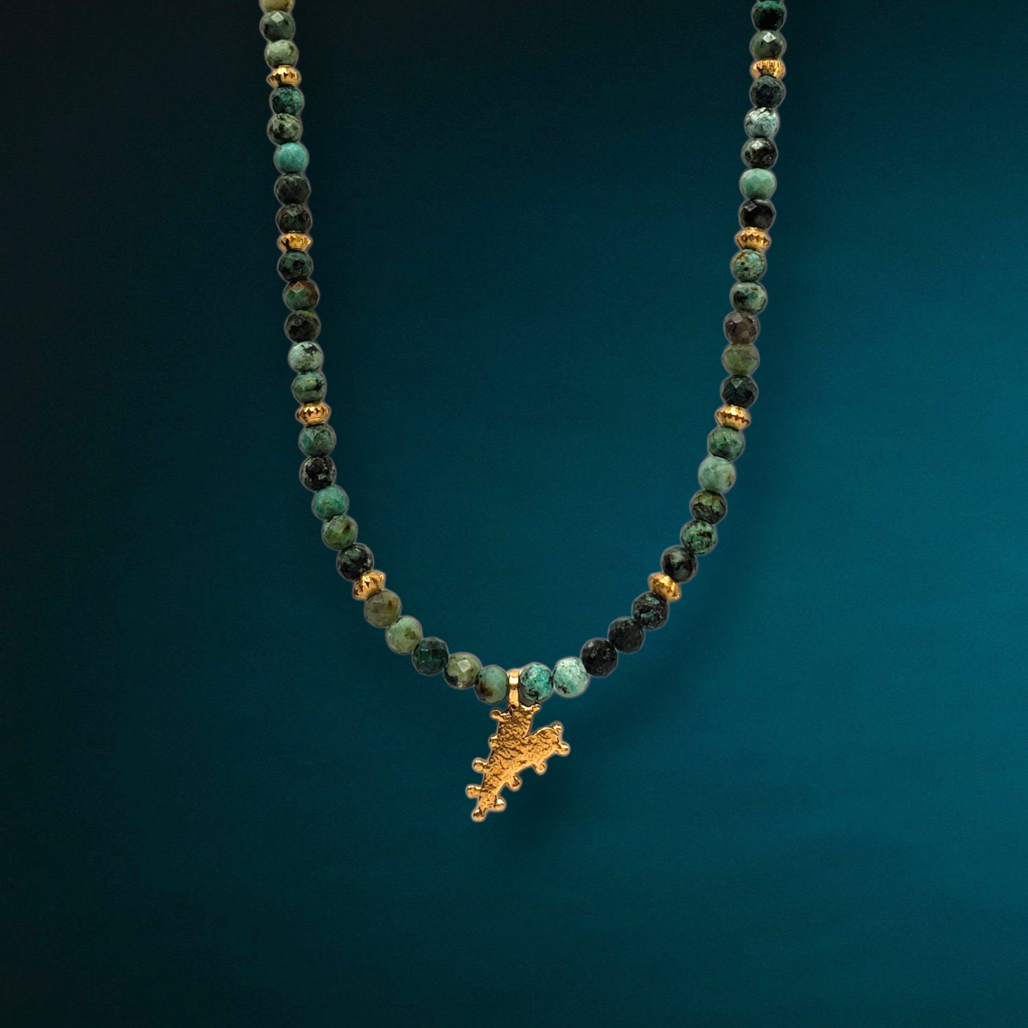 The Chrysocolla Beaded Heart Charm Necklace by Dreamers of Dreams features green and black marbled beads, gold accents, and a heart charm, capturing bohemian style against a gradient teal backdrop.