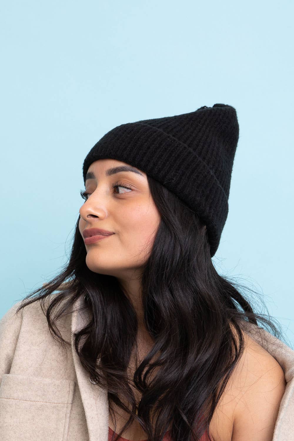 A woman with long dark hair wears the Leto Accessories Versatile Cuffed Beanie, a cozy rib knit hat, along with a beige coat, looking to the side against a light blue background.