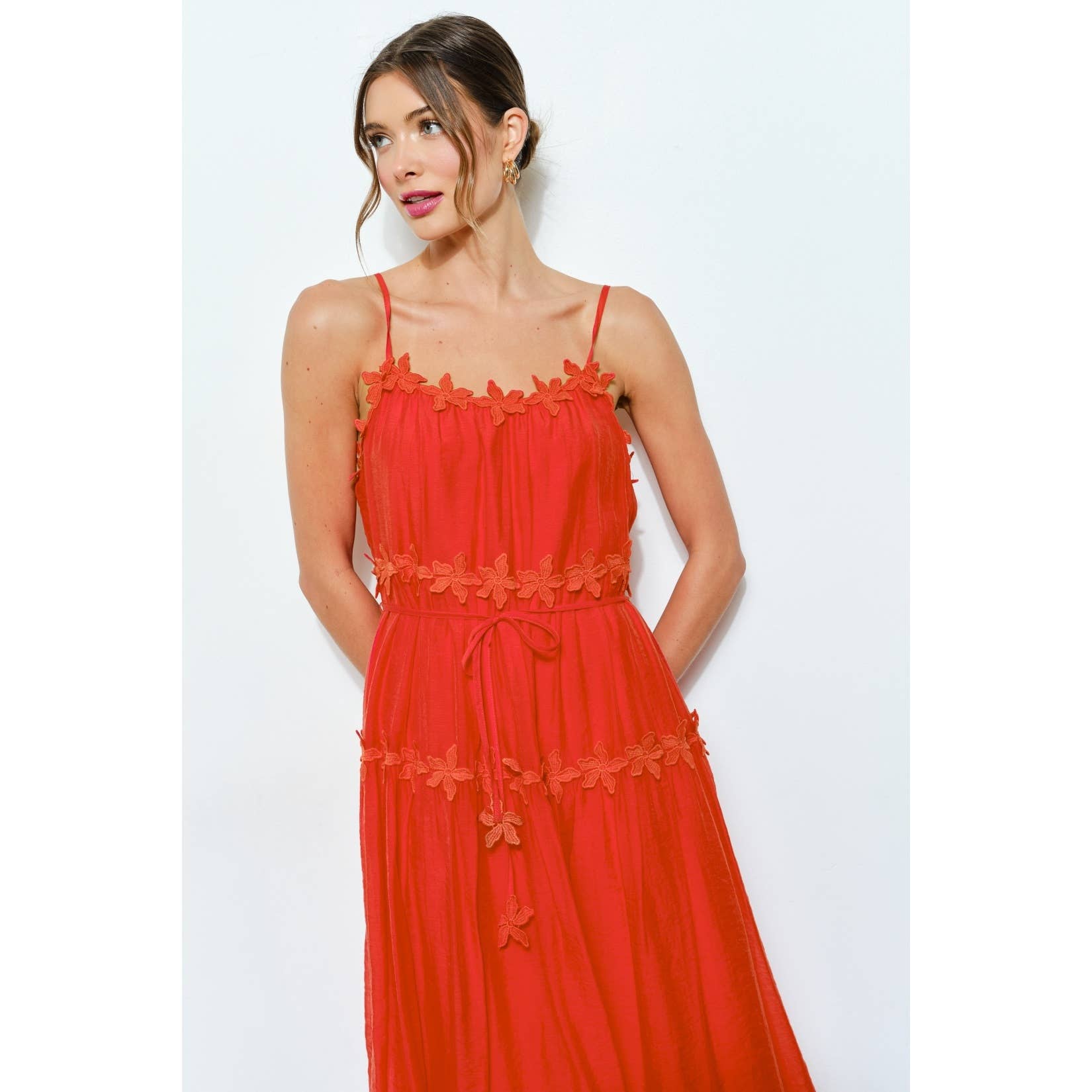 A woman in the INA Sleeveless Floral Applique Tiered Midi Dress, featuring thin straps and floral accents at the neckline, waist, and skirt, stands against a plain white backdrop with her hair in a loose updo.